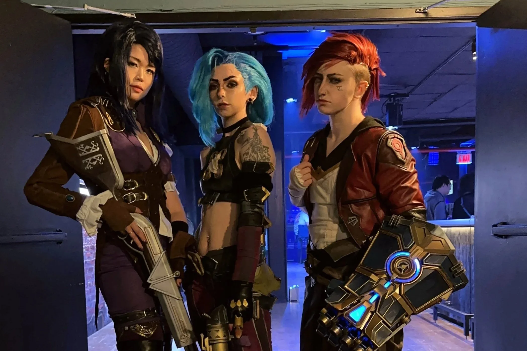 Three people dressed as characters from a video game, with elaborate costumes and weapons, posing indoors at an event.