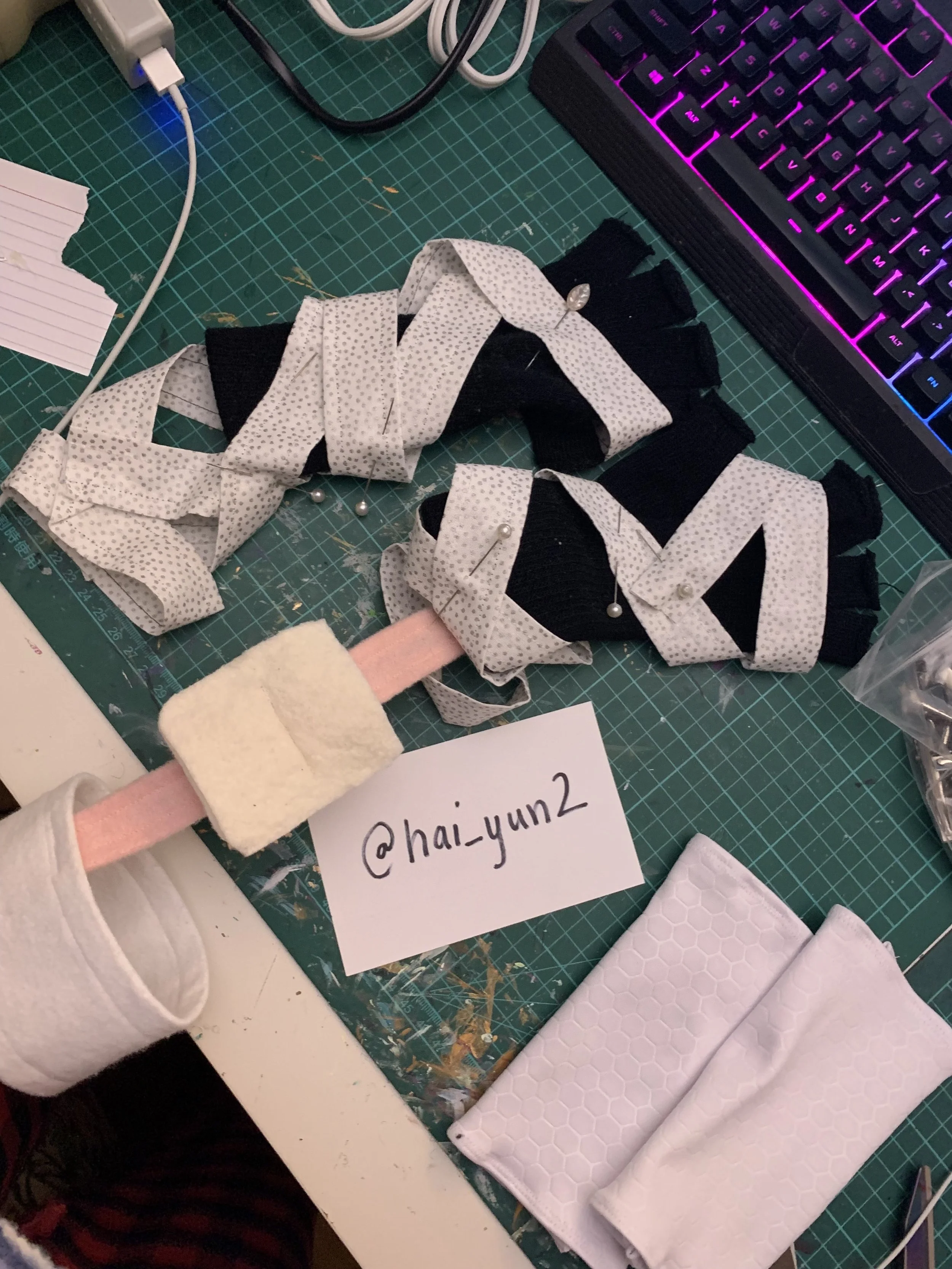 A cluttered craft workspace with fabric, sewing pins, and a partially sewn fabric piece. There is a sewing rollback with a pink strap and attached felt pad, a white piece of fabric with a honeycomb pattern, and a computer keyboard with colorful backl