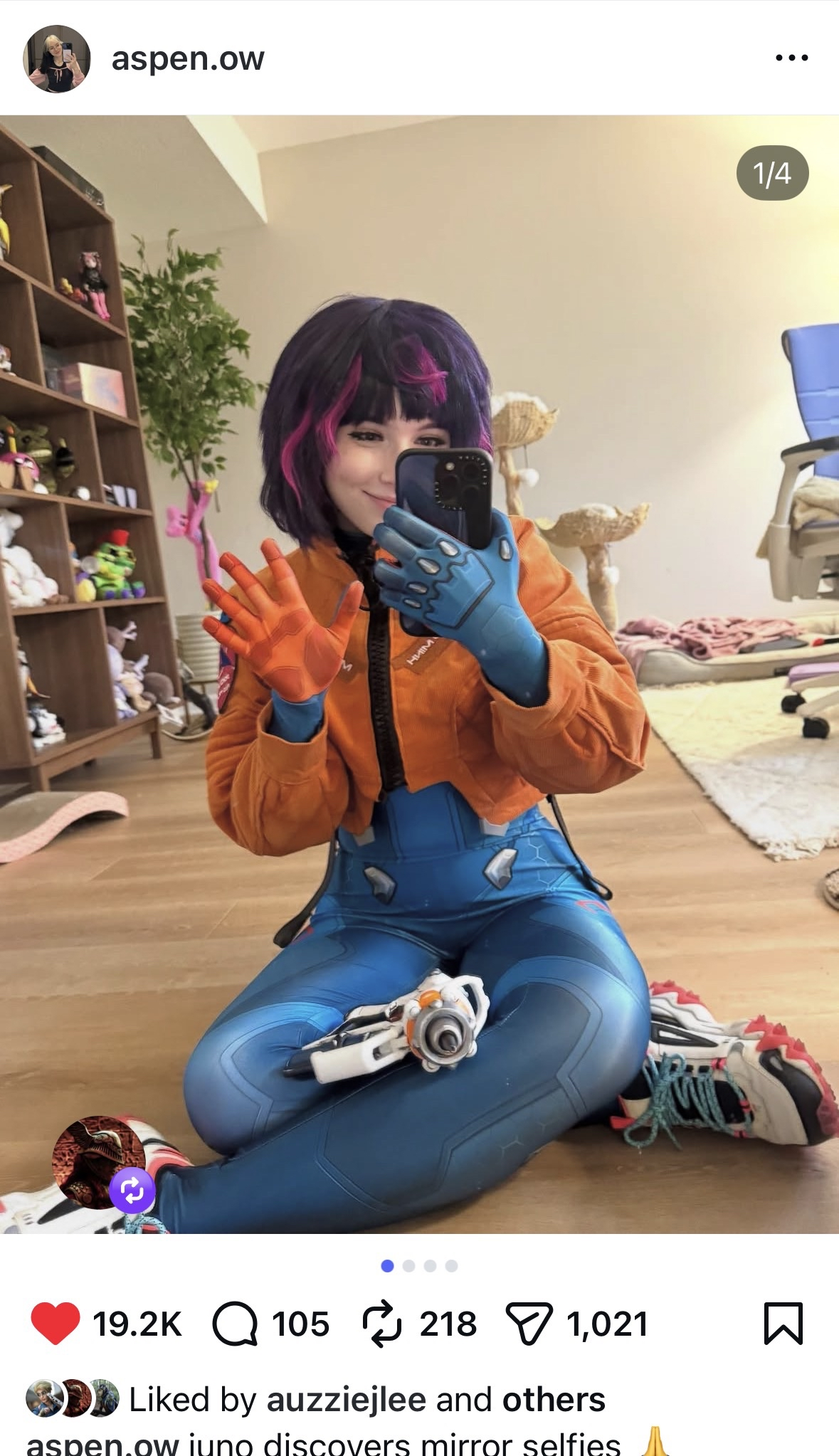 A woman with purple and pink hair taking a mirror selfie in a room. She is wearing a costume with a blue robotic arm and gloves, an orange jacket, and blue pants. She is sitting on the floor, smiling, with a collection of plush toys and objects in th