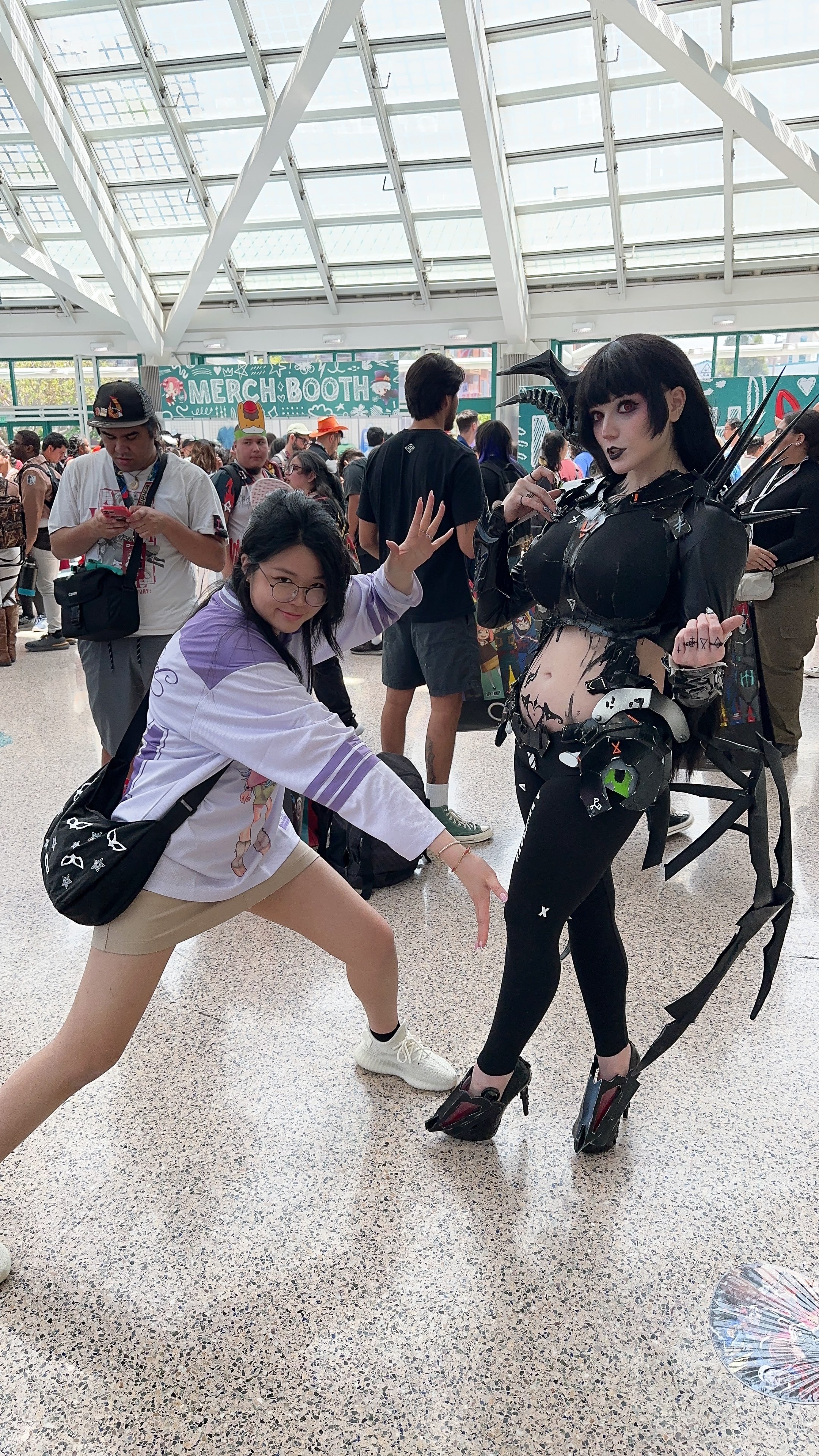 A woman in elaborate cosplay with black hair, horns, dark makeup, and a black costume stands next to a woman in casual clothing at a convention. Other attendees are in the background, with a sign that reads 'MERCH BOOTH' visible in the indoor setting