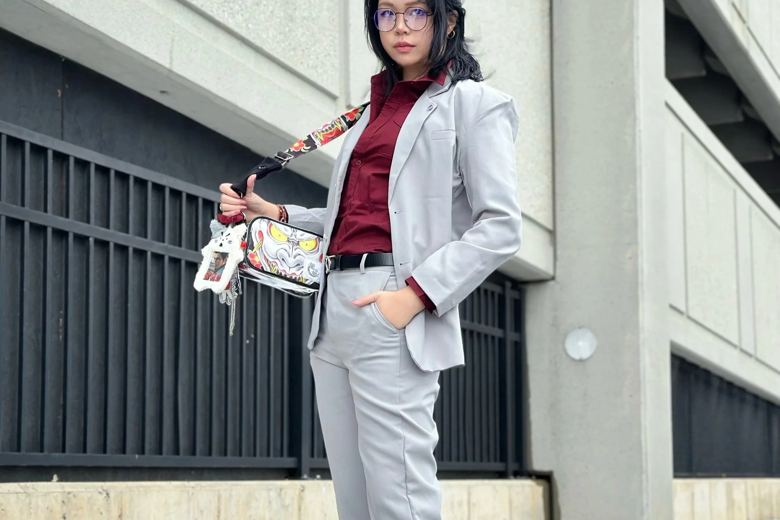 A woman wearing a light gray suit with a red shirt, glasses, and holding a colorful umbrella and purse, standing outdoors near a modern building with concrete walls and black railing.