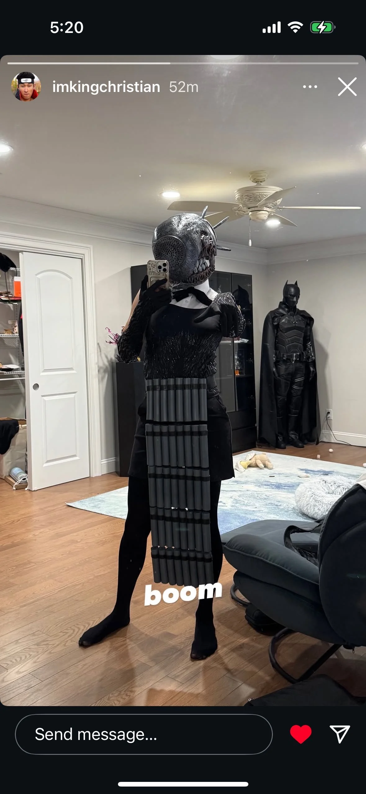 Person in a costume with a skull mask taking a selfie in a mirror, standing in a room with Batman costume in the background.