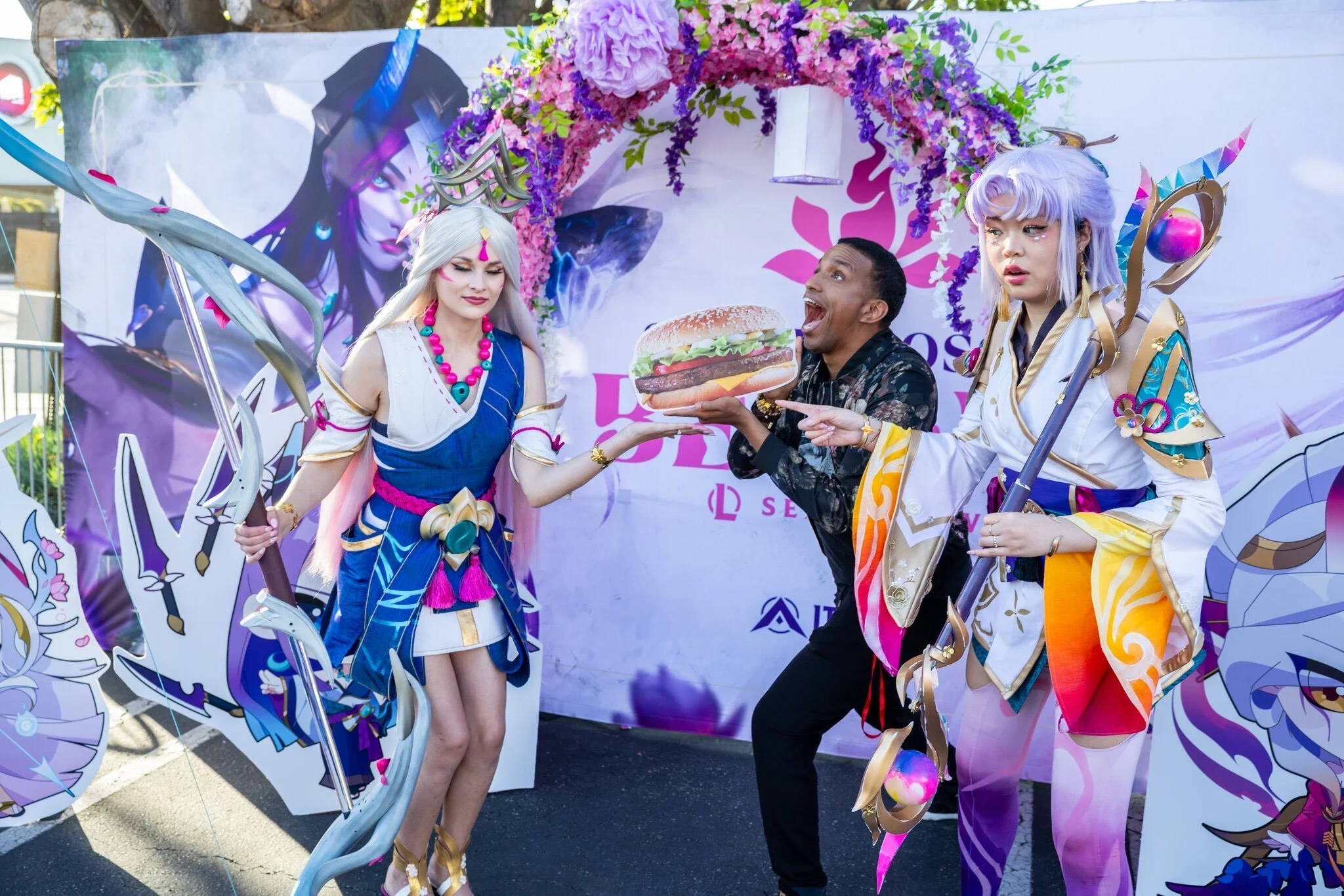 Two women dressed in elaborate fantasy costumes with weapons and accessories, one holding a large paper burger, standing beside a man holding the burger and pointing at it, with a fantasy-themed backdrop featuring characters and decorative flowers.