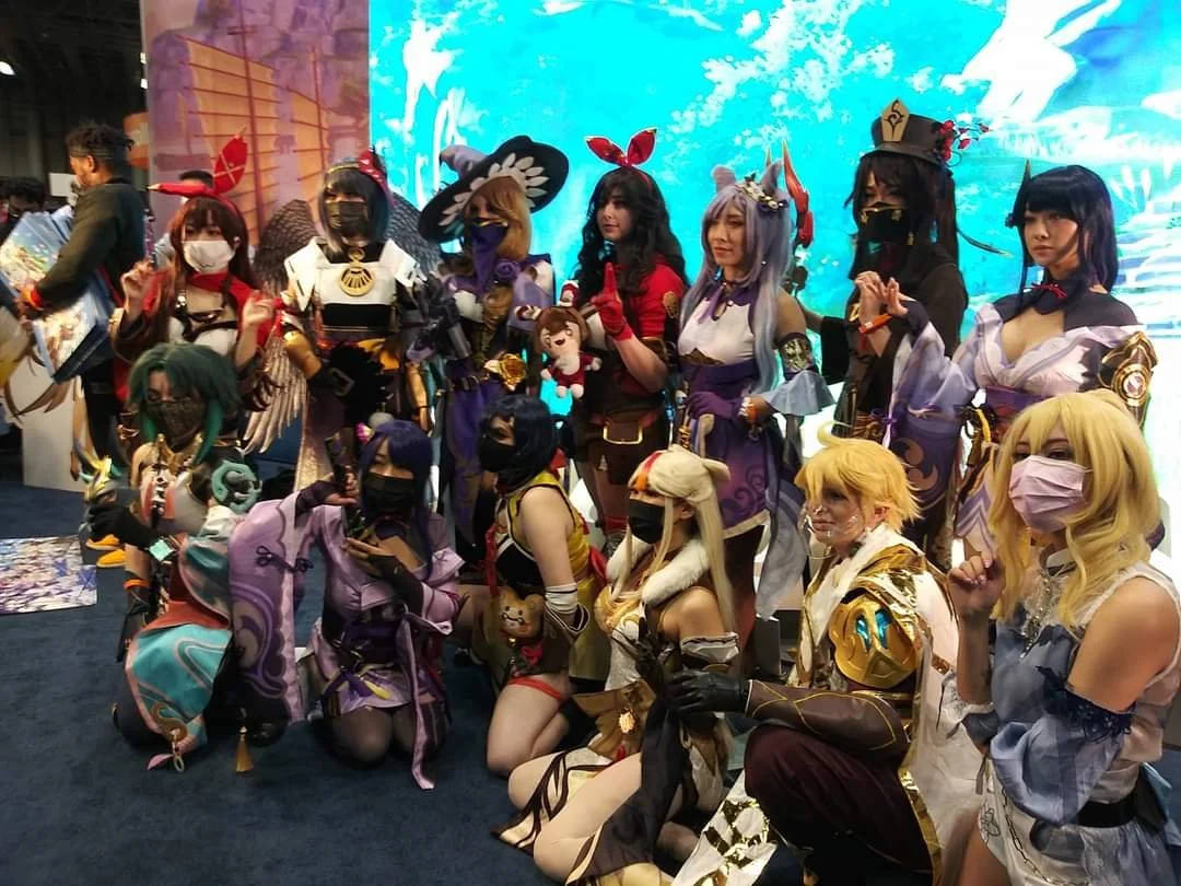 Group of people dressed as characters from a video game or anime, in elaborate costumes, at a convention. Some are kneeling in front, others standing behind, with a digitally animated background.