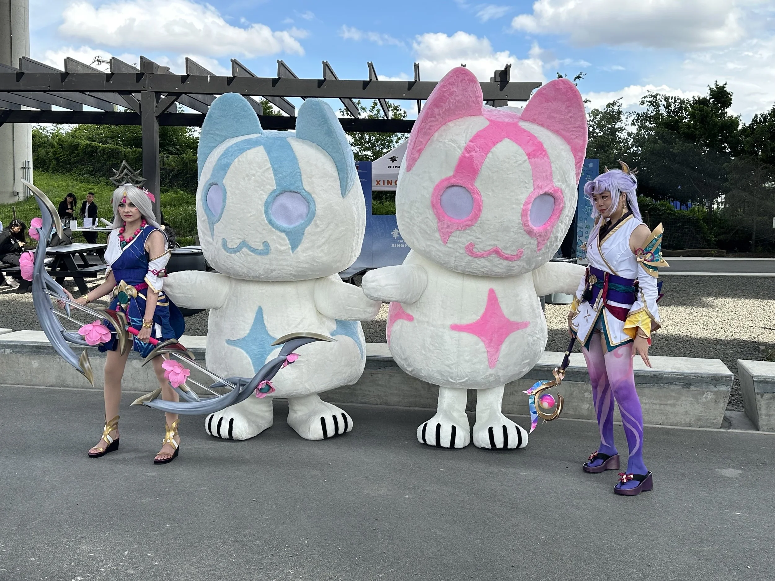 Two women in cosplay costumes posing with large mascot characters, one blue and one pink, at an outdoor event.
