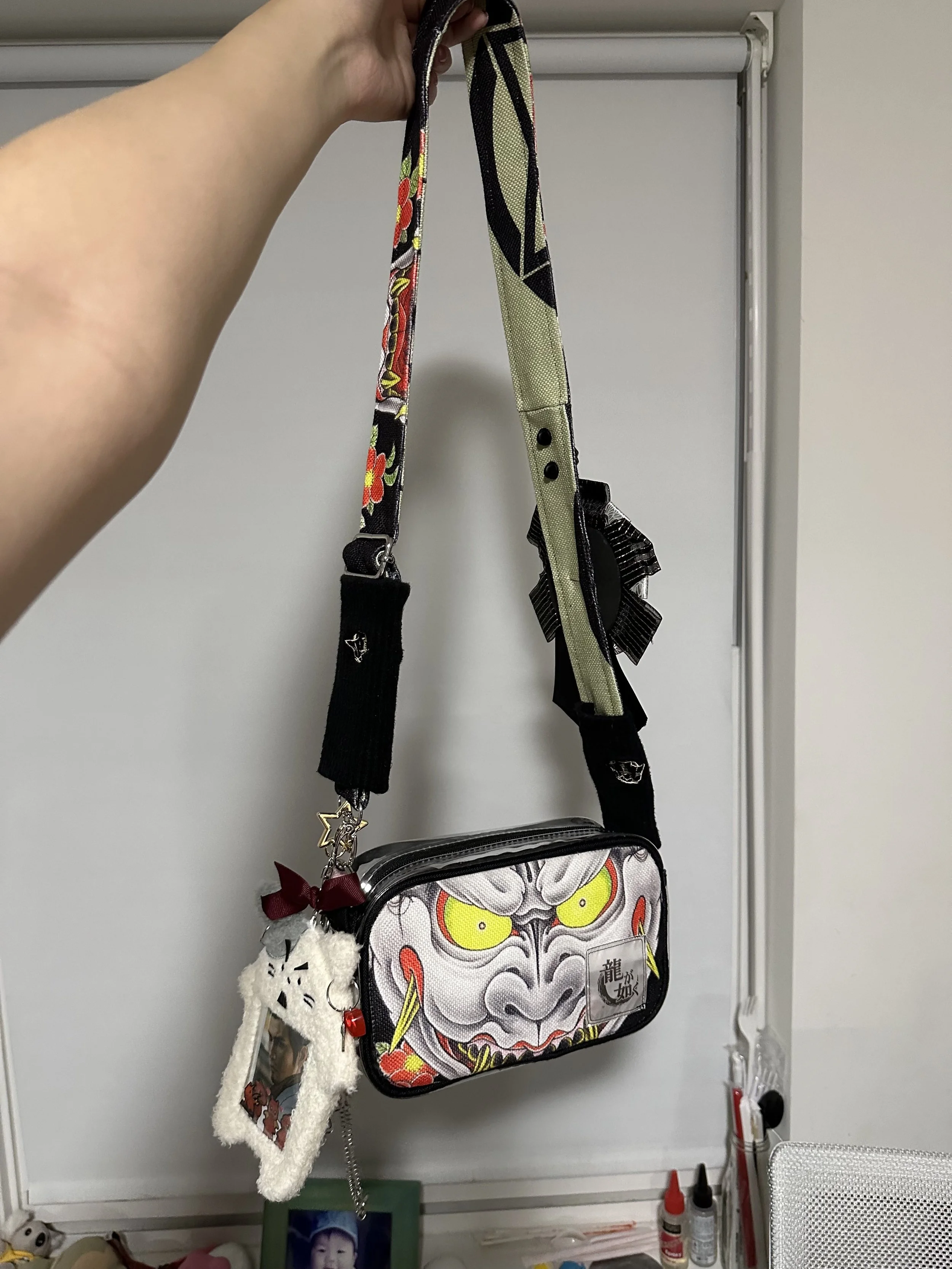 A colorful handbag with an Oni mask face design hanging from a hand, with plush keychains attached, in front of a whiteboard and desk with bottles and photo frames.