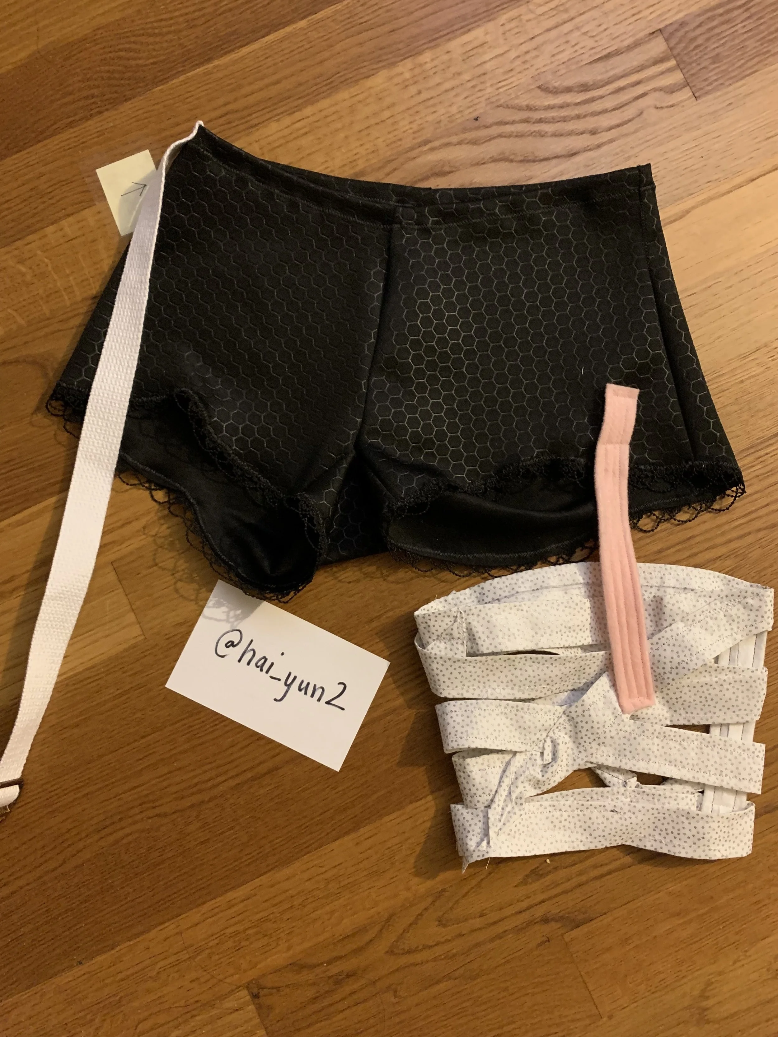 Black lace boyshort underwear, pink garter, and white folded fabric with small black dots on a wooden surface, with a handwritten note '@hai_yun2'.