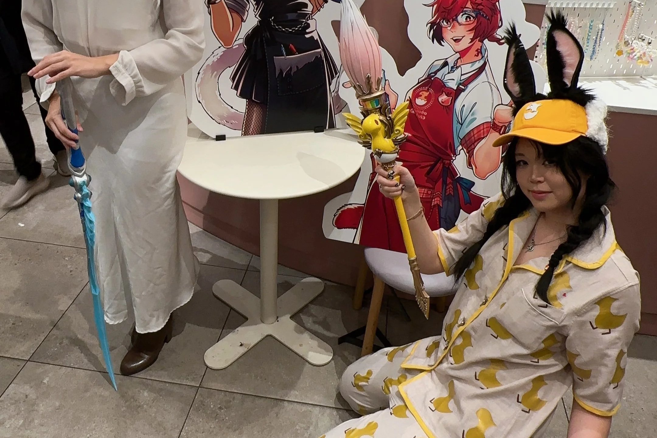 A woman dressed in pajamas with duck prints, wearing a yellow hat with bunny ears, is sitting on the floor holding a staff with a yellow duck figure. She is at a convention or event, with a colorful anime-style poster behind her. There is a partially visible person next to her holding a blue umbrella, and a paper cut-out of an anime character in the background.