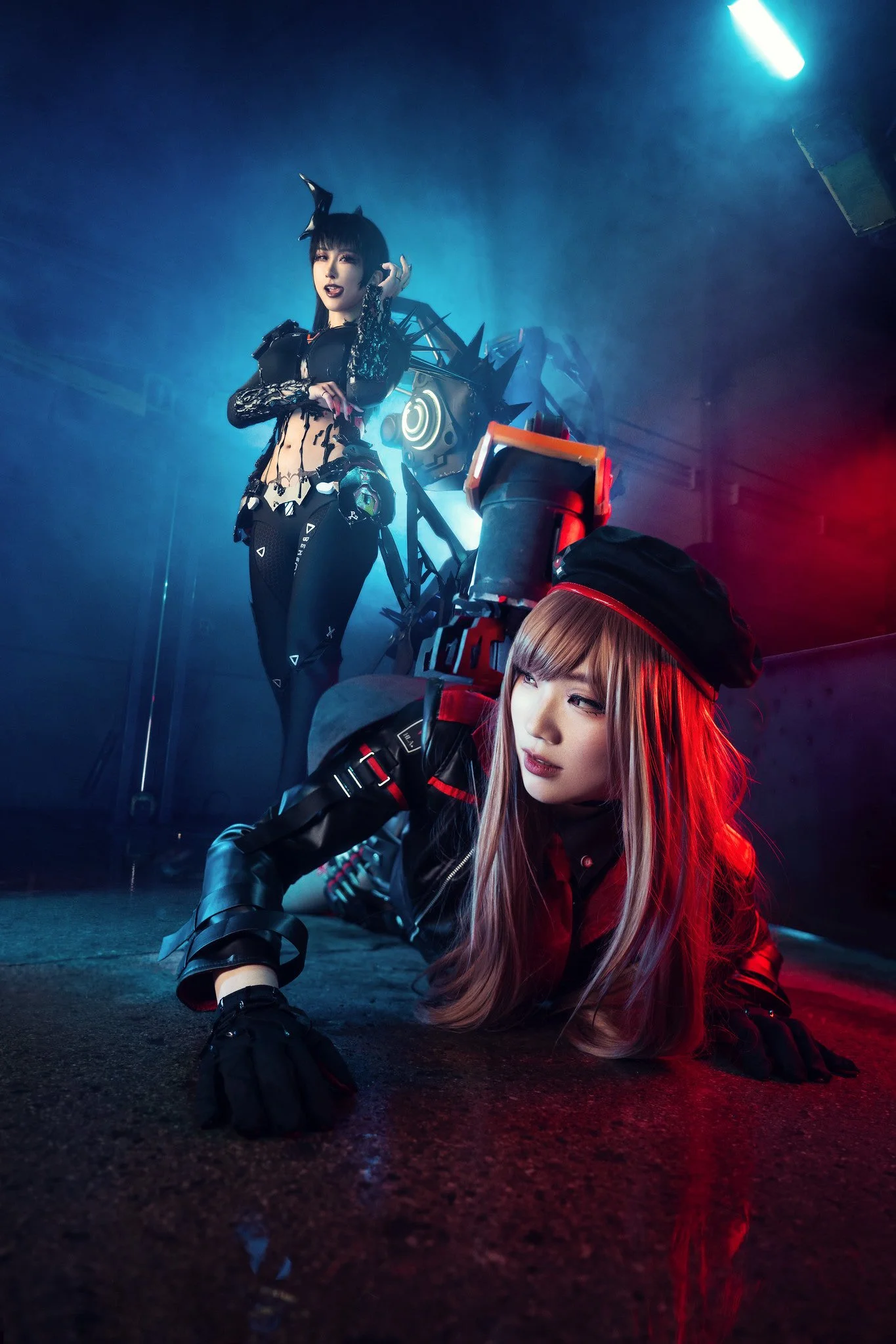 Two women in elaborate costumes with cyberpunk and fantasy elements on a dark stage, one kneeling on the ground and the other standing behind her.