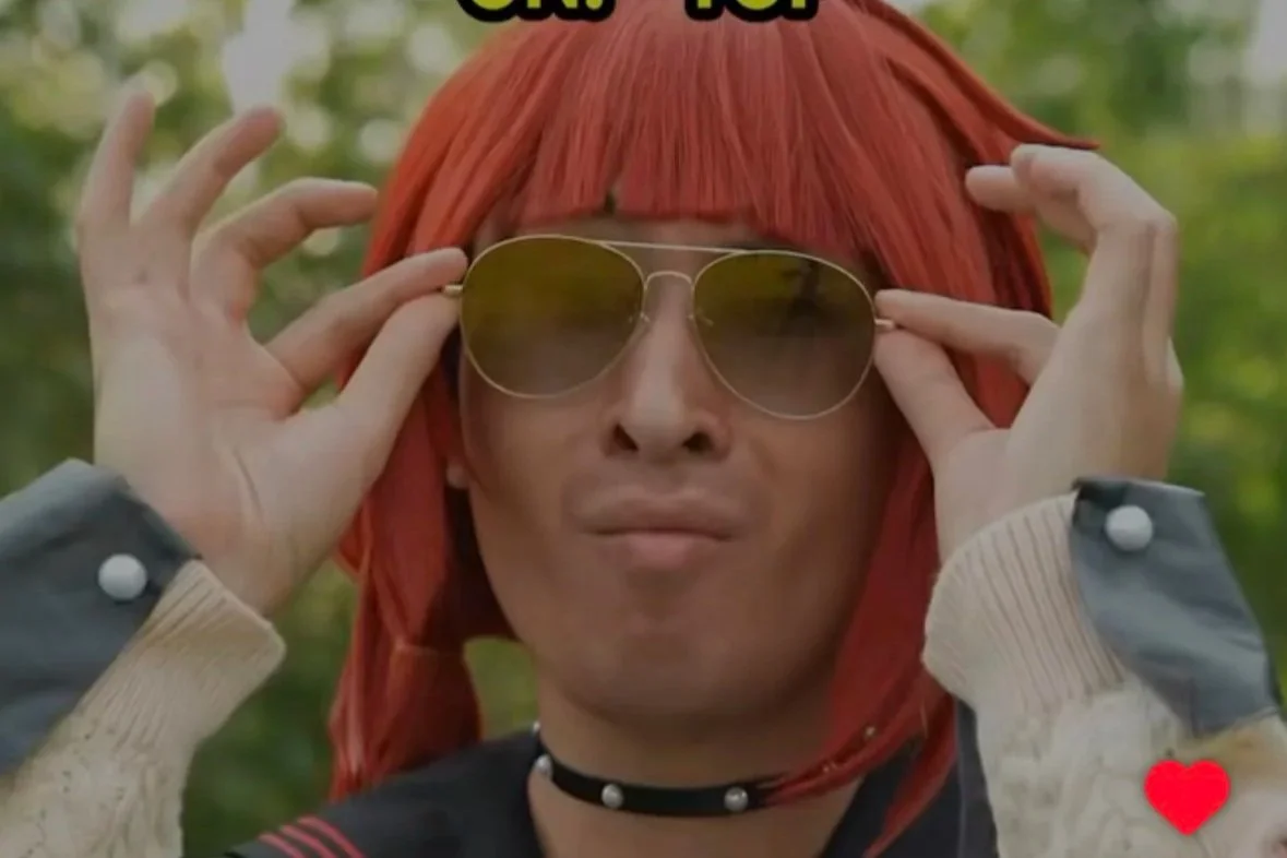 Person with red hair and sunglasses adjusting them outdoors, wearing a choker necklace and a jacket with striped cuffs, with a blurred green background.