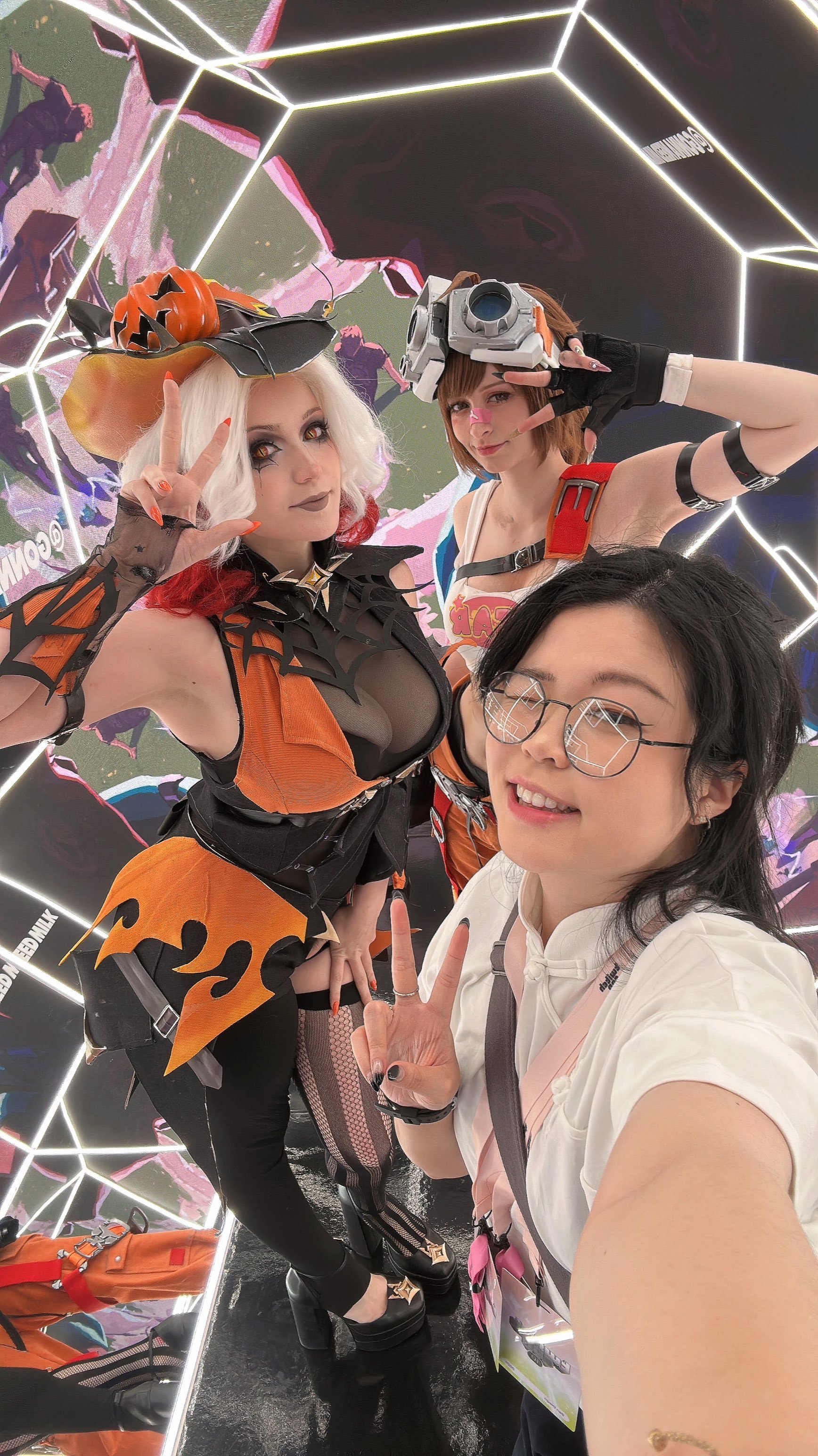 Three women in cosplay taking a selfie at a convention or event, with Hispanic and Asian features, two of whom are in Halloween-themed costumes. The woman on the left has white hair with red tips, a Halloween hat, and a black and orange costume with 
