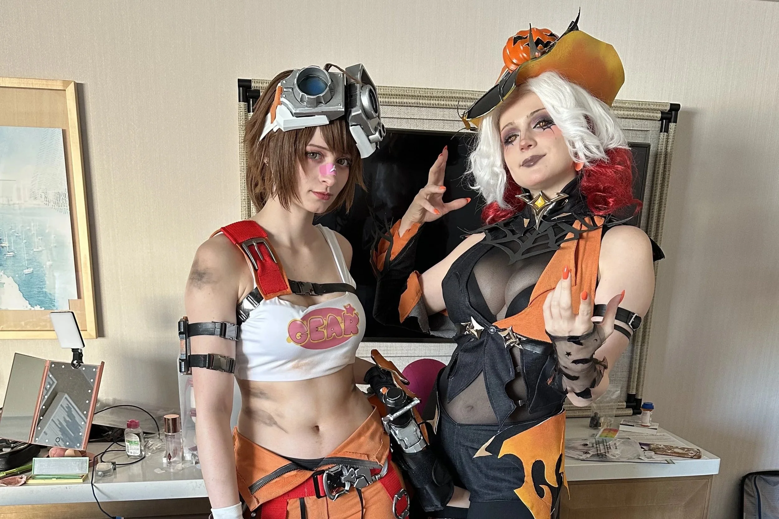 Two women in detailed Halloween costumes posing indoors with a plain wall and framed picture in the background. The woman on the left has short brown hair, wears a white crop top with "GEAR" on it, orange and black shorts, and a large robotic headpiece. The woman on the right has white and red hair, a pumpkin hat, black mesh top under an orange and black costume, and black gloves with orange nails.