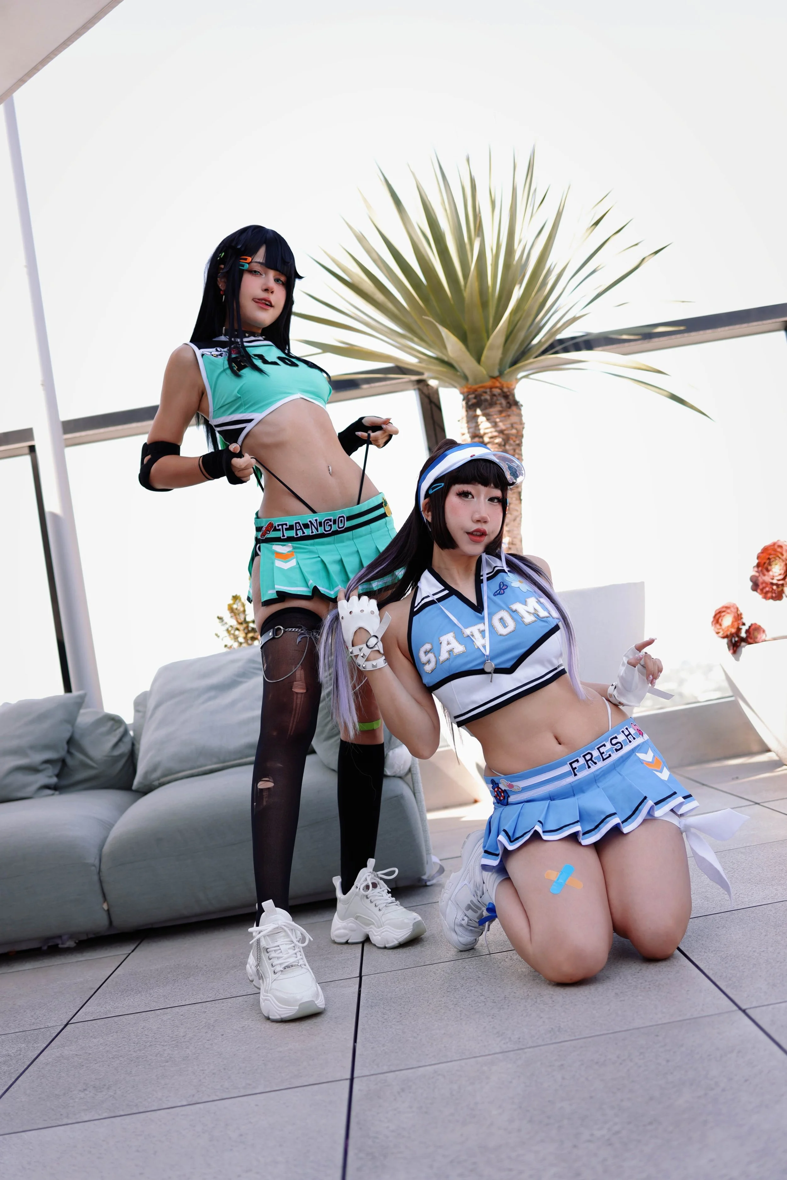 Two young women dressed in colorful, sporty cosplay outfits posing outdoors on a tiled patio with a gray sofa, a palm tree, and a white canopy in the background.