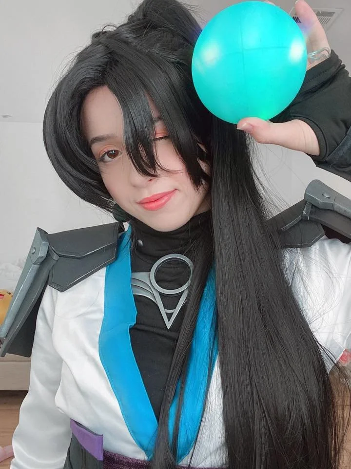 A woman in a cosplay costume holding a blue ball, winking, with black hair styled in a theme-related outfit.
