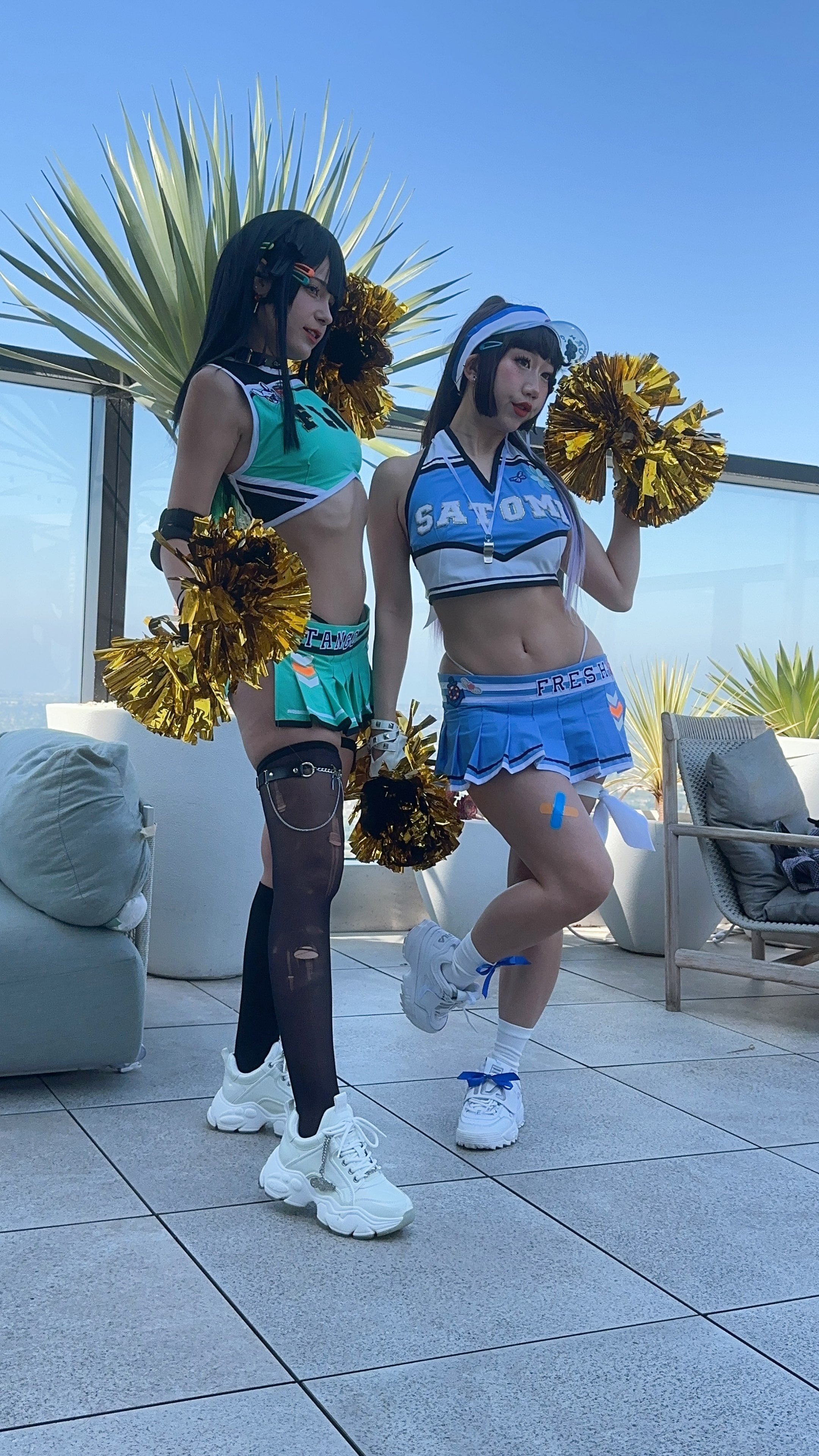 Two women dressed as cheerleaders with pom-poms posing outdoors on a rooftop patio with a city view, wearing colorful mini skirts and crop tops.