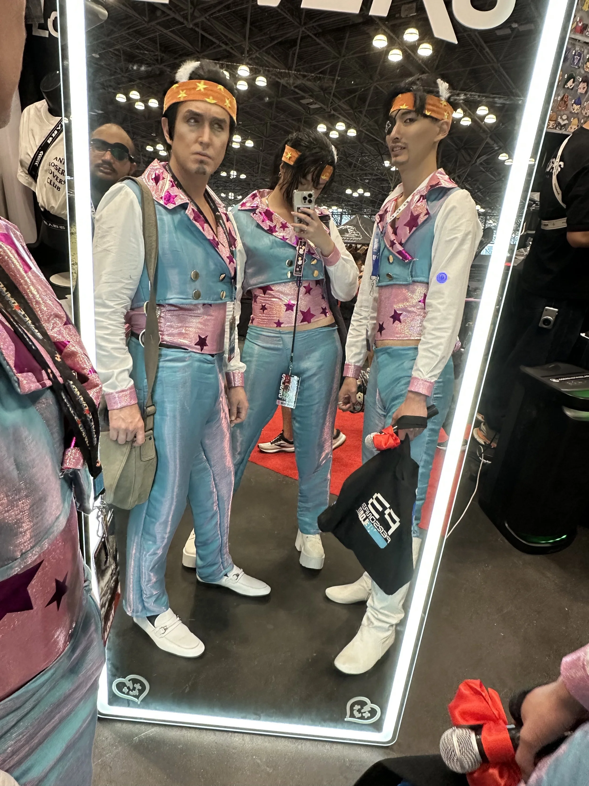 Three people in colorful, shiny outfits with stars, taking a mirror selfie at a convention with a person in the background wearing sunglasses.