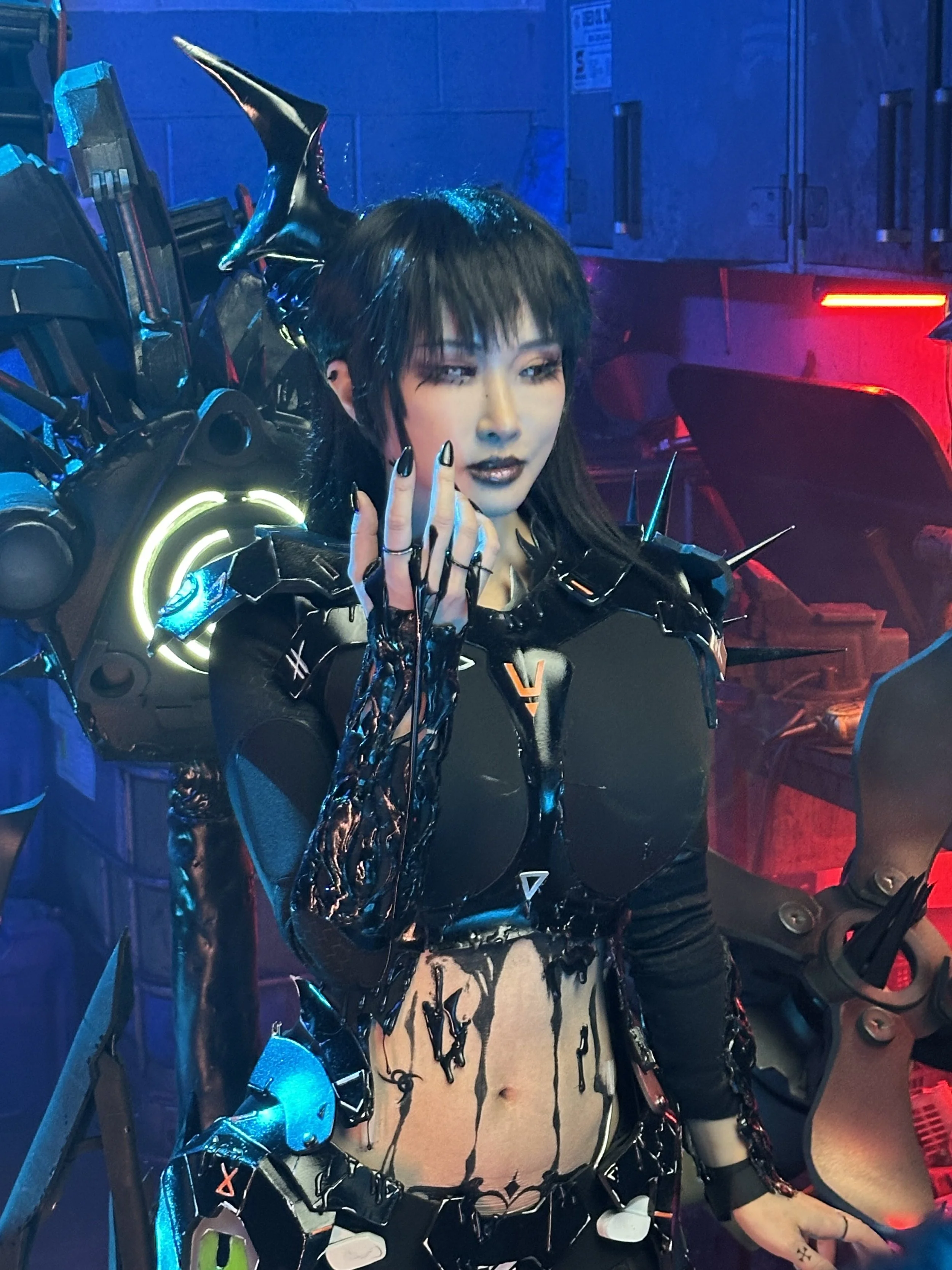 A woman in a futuristic, cyberpunk-style costume with black and metallic elements, shiny black makeup, and a headpiece with sharp, wing-like protrusions, standing in front of sci-fi props and equipment with blue and red lighting.