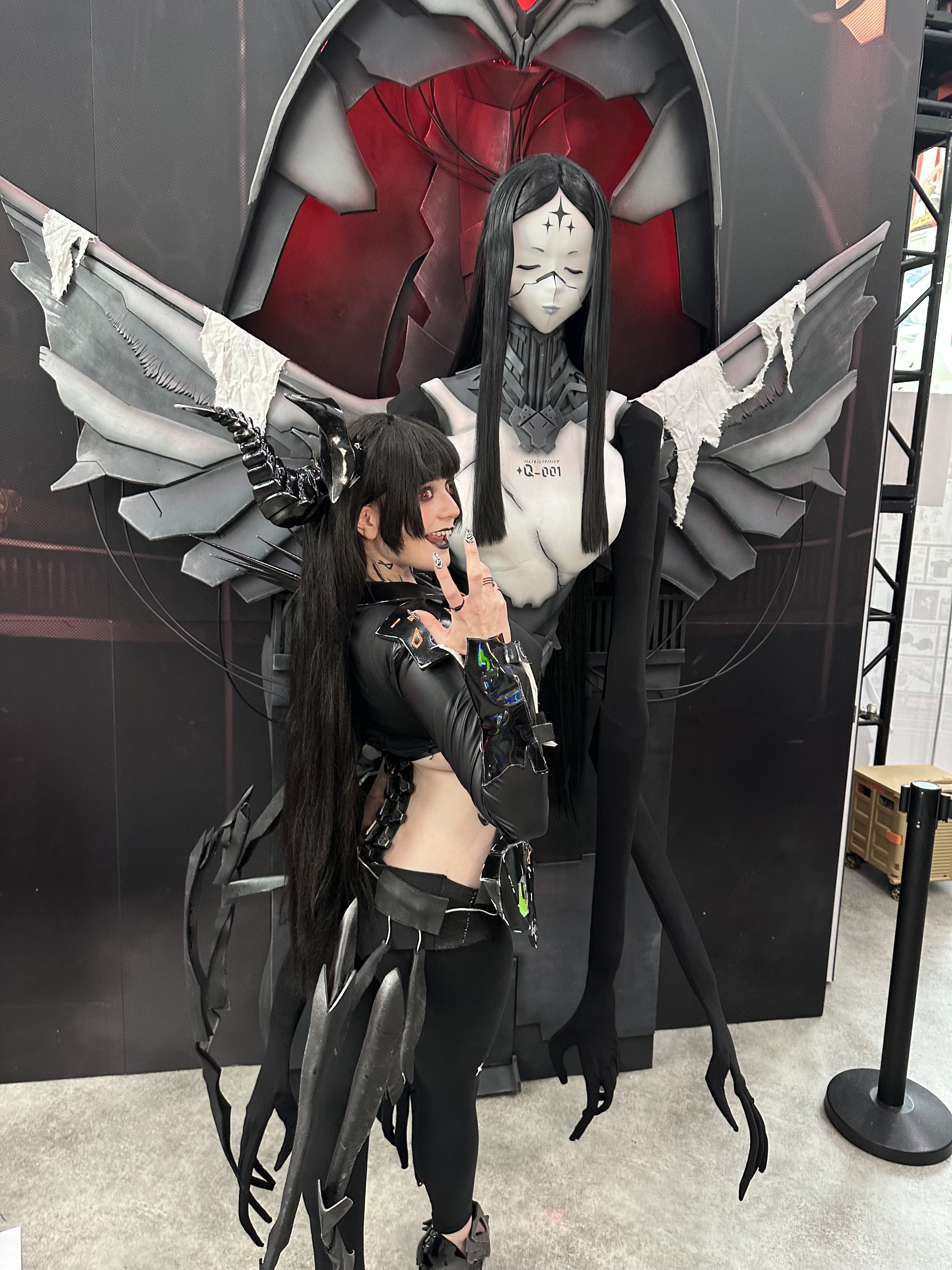 Person in a black, sci-fi inspired costume posing near a large, robotic figure with a feminine, anime-style face and elaborate wings.