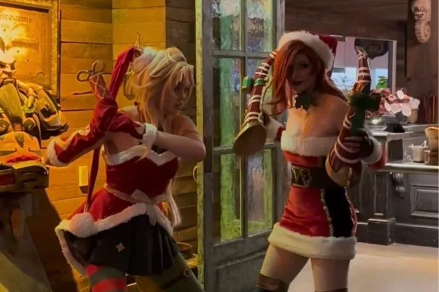 Two women dressed in Christmas-themed costumes posing indoors, with one dressed as Santa and the other in a festive outfit with reindeer antlers, in a cozy holiday setting.