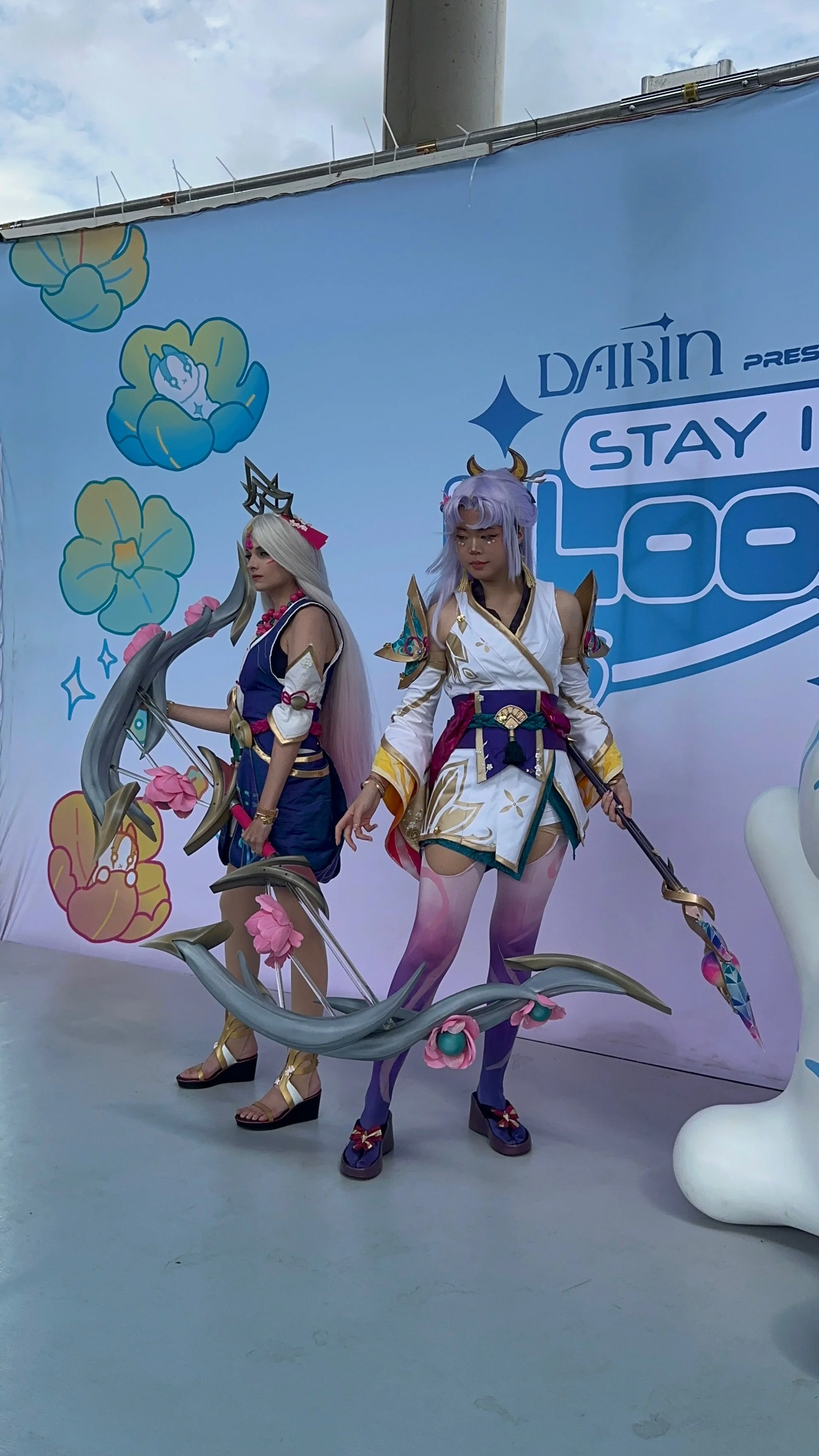 Two women in cosplay costumes posing in front of a backdrop with a blue and white design and the text "DABIN presents STAY COOL." They are dressed as characters with elaborate costumes, including accessories, wigs, and makeup, with a large decorative prop resembling a slithering creature with pink flowers.