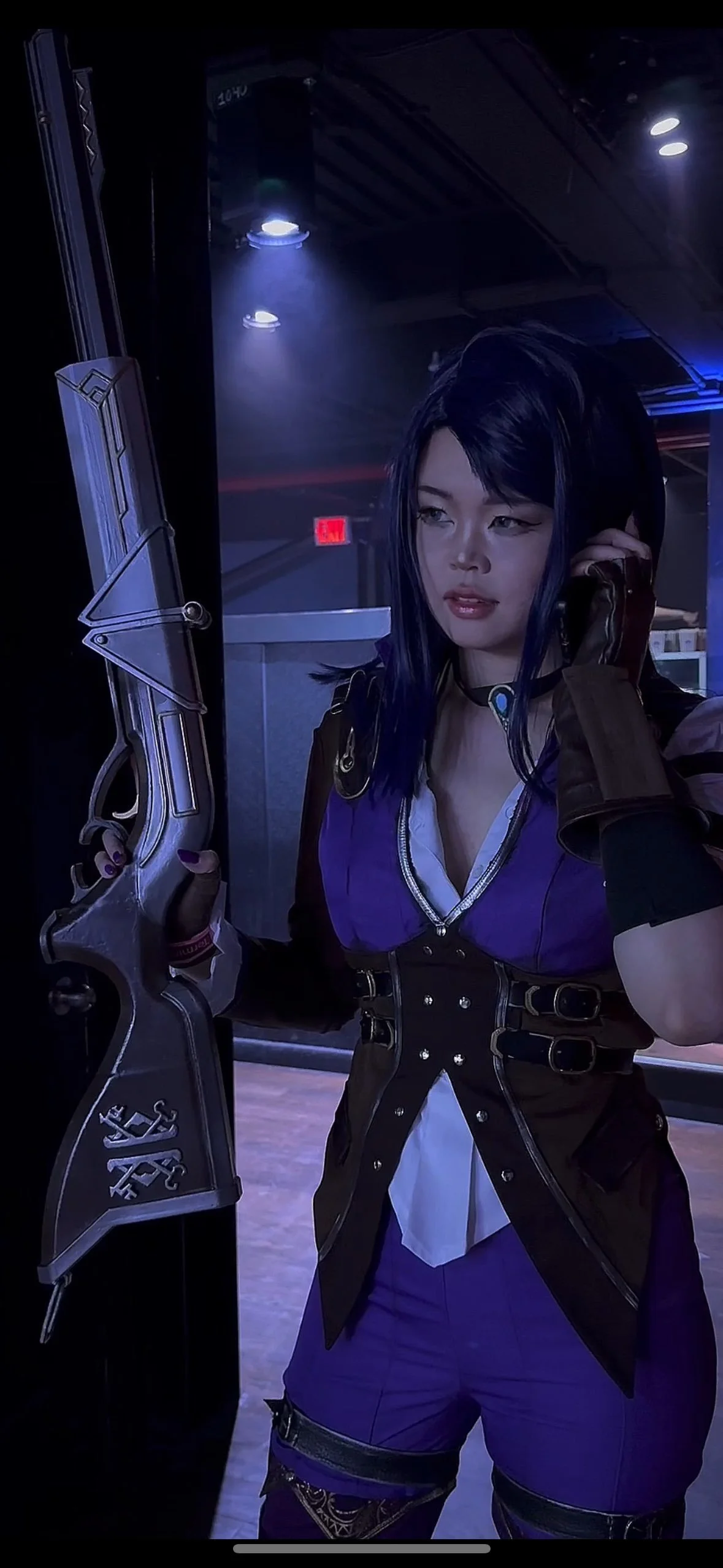A woman with dark hair in a costume, holding a futuristic-looking rifle, talking on a phone in a dimly lit room with blue lighting.
