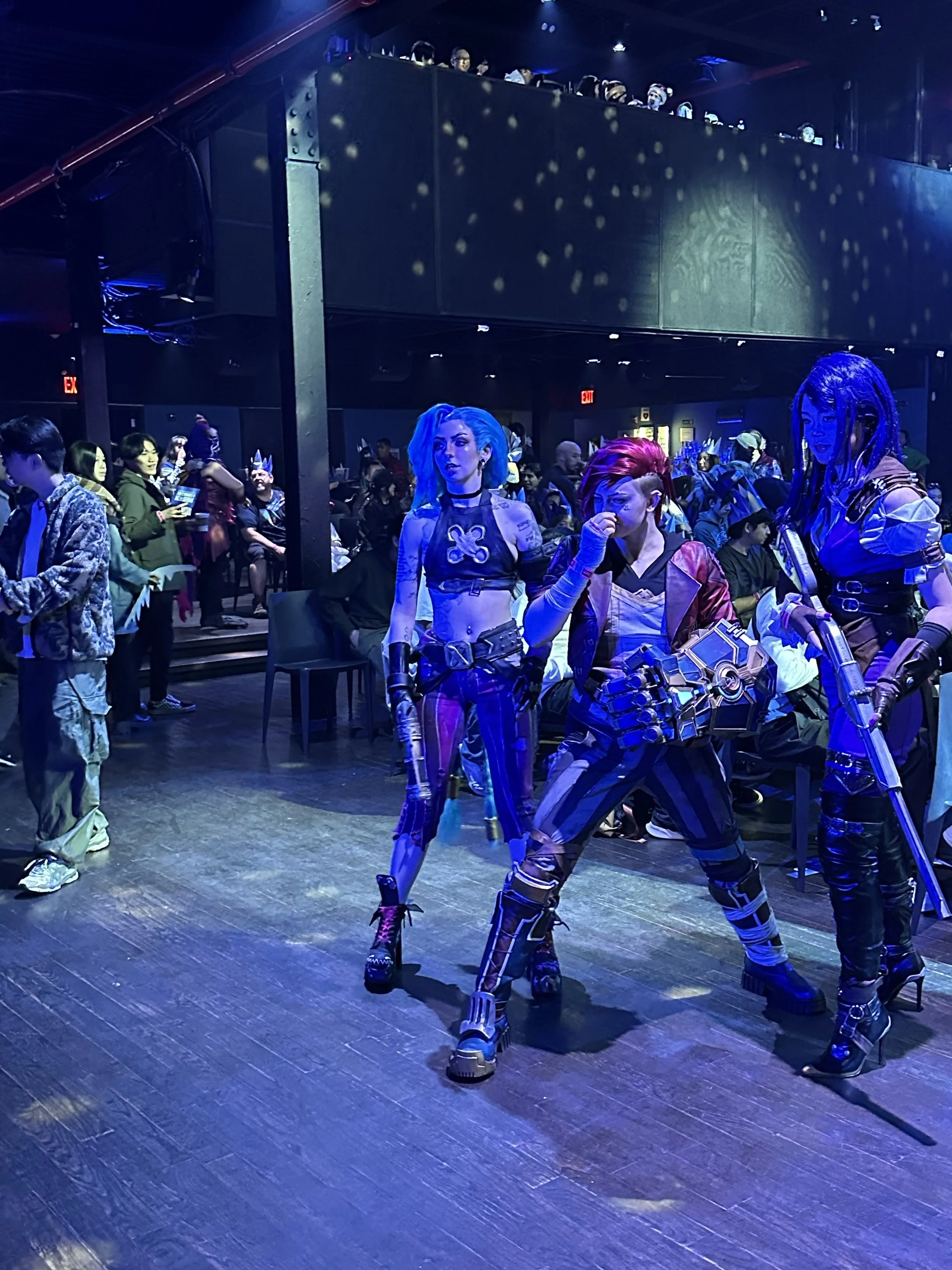 Three women dressed in futuristic, cyberpunk costumes pose on a dark, illuminated stage at a convention. The background is filled with seated and standing attendees, some in costume, under blue and purple lighting.
