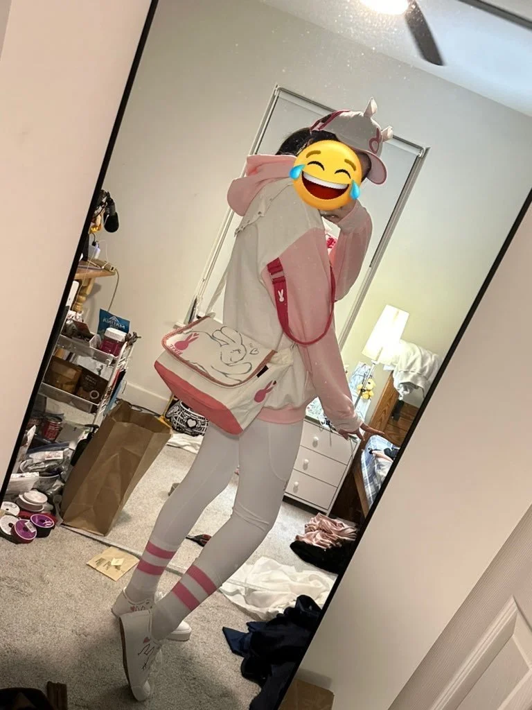 Person taking a mirror selfie in a bedroom wearing a pink and white hoodie, white leggings with pink stripes, and white shoes, with a cartoon-themed bag.