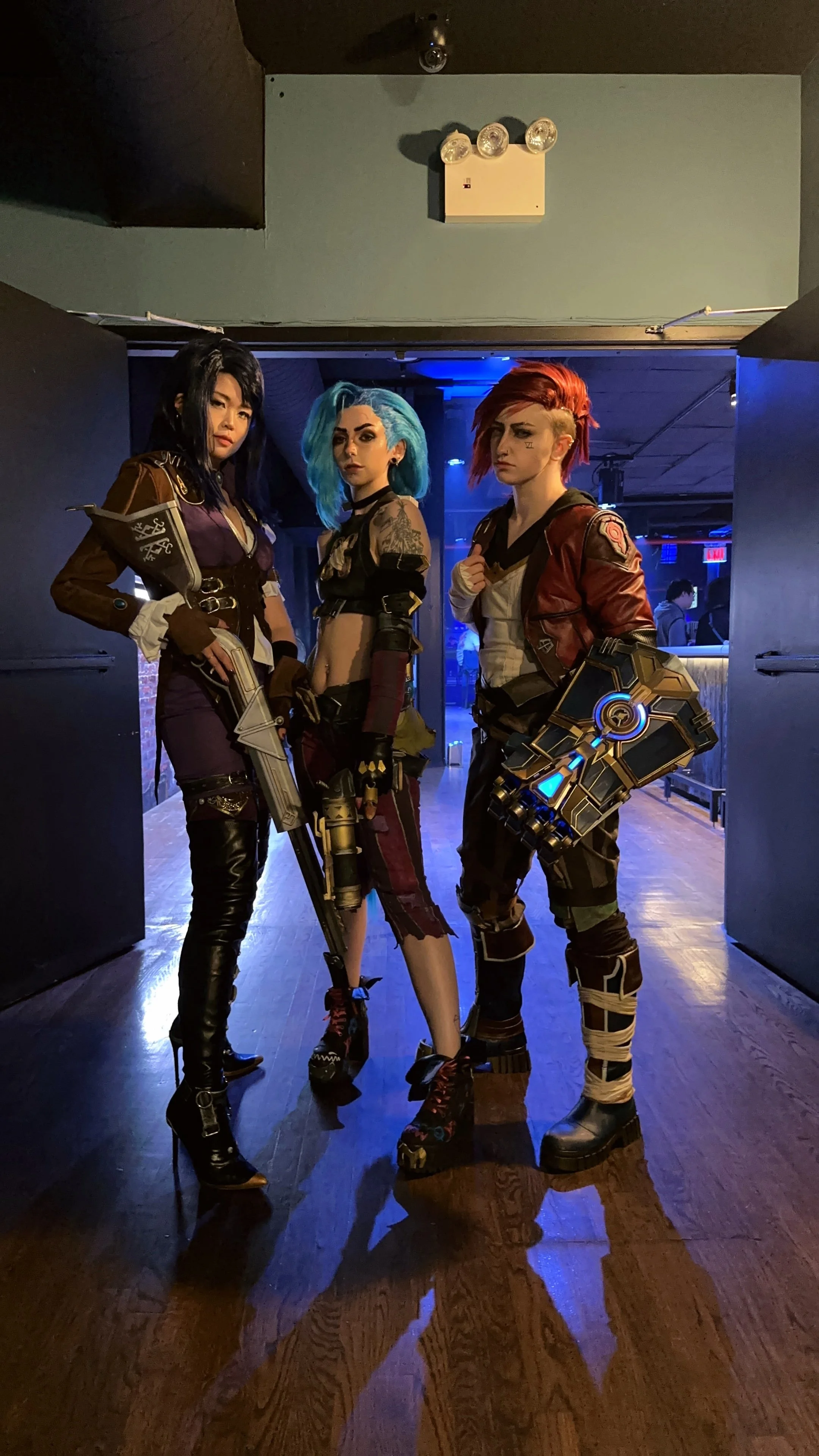 Three people dressed in cosplay costumes at an indoor event, standing on a wooden floor with a dark background and blue lighting.