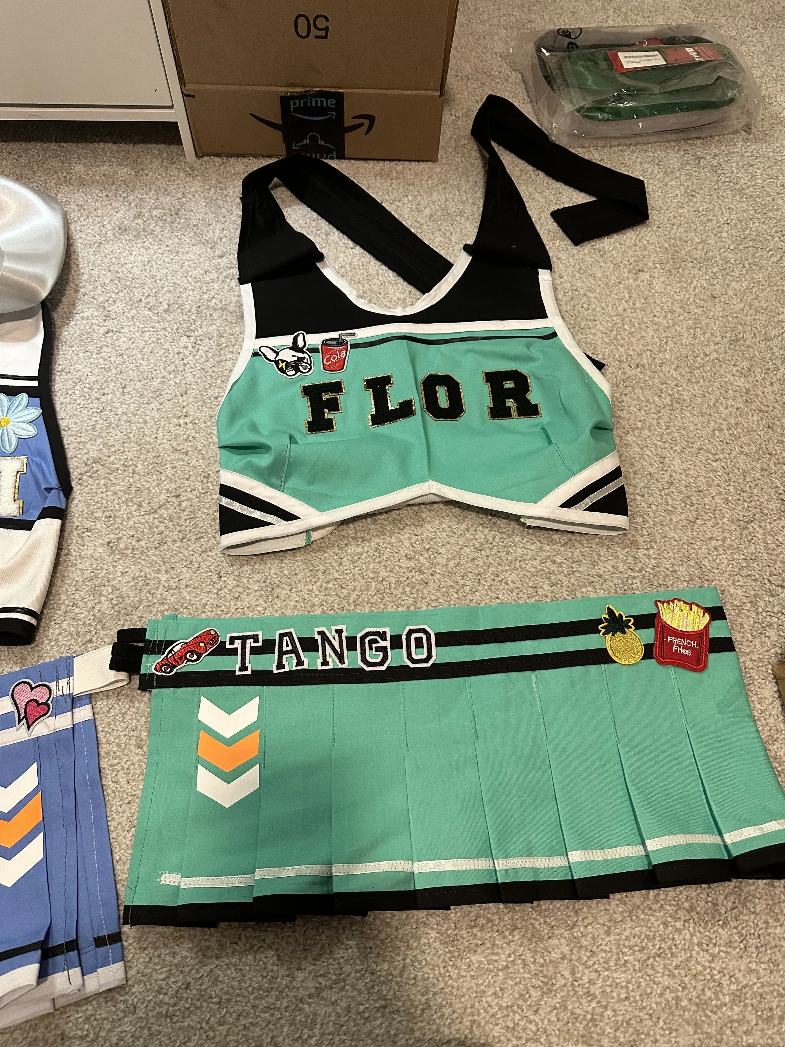 A cheerleading outfit with a black and teal top embroidered with the word 'FLOR' and patches, and a matching teal skirt with patches and the word 'TANGO' on the waistband, laid out on beige carpet.