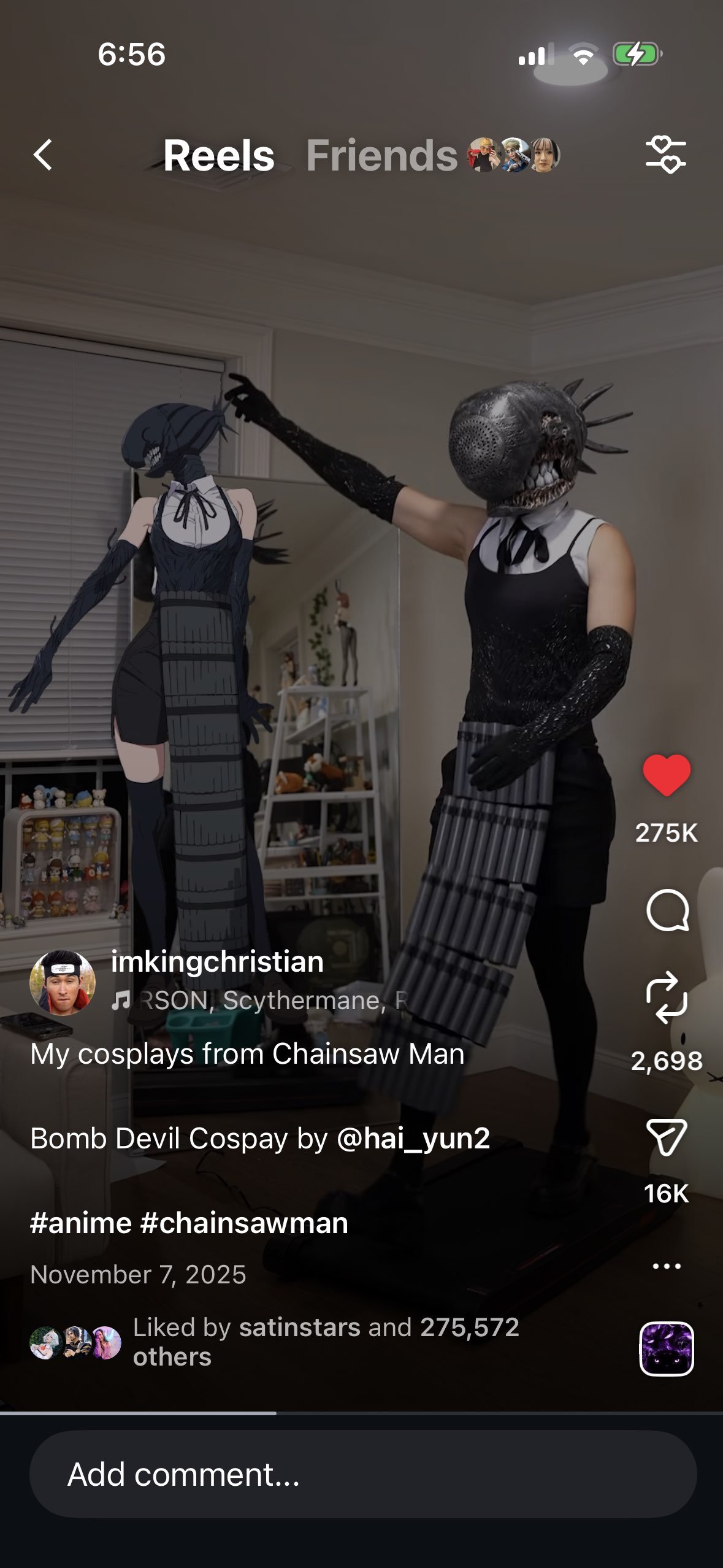 Person wearing a mask with an alien-like face and a dress, pointing at a life-sized anime character cutout of a girl with blue hair, black dress, and stockings, in a living room.