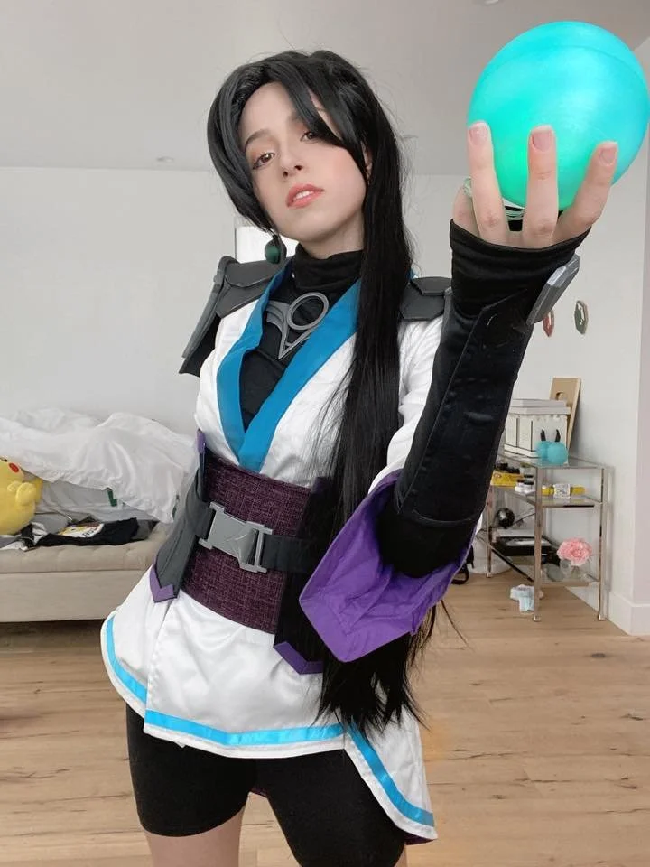 Person in elaborate costume holding glowing blue orb in hand, standing in a room with wooden floor, bed, and shelves.