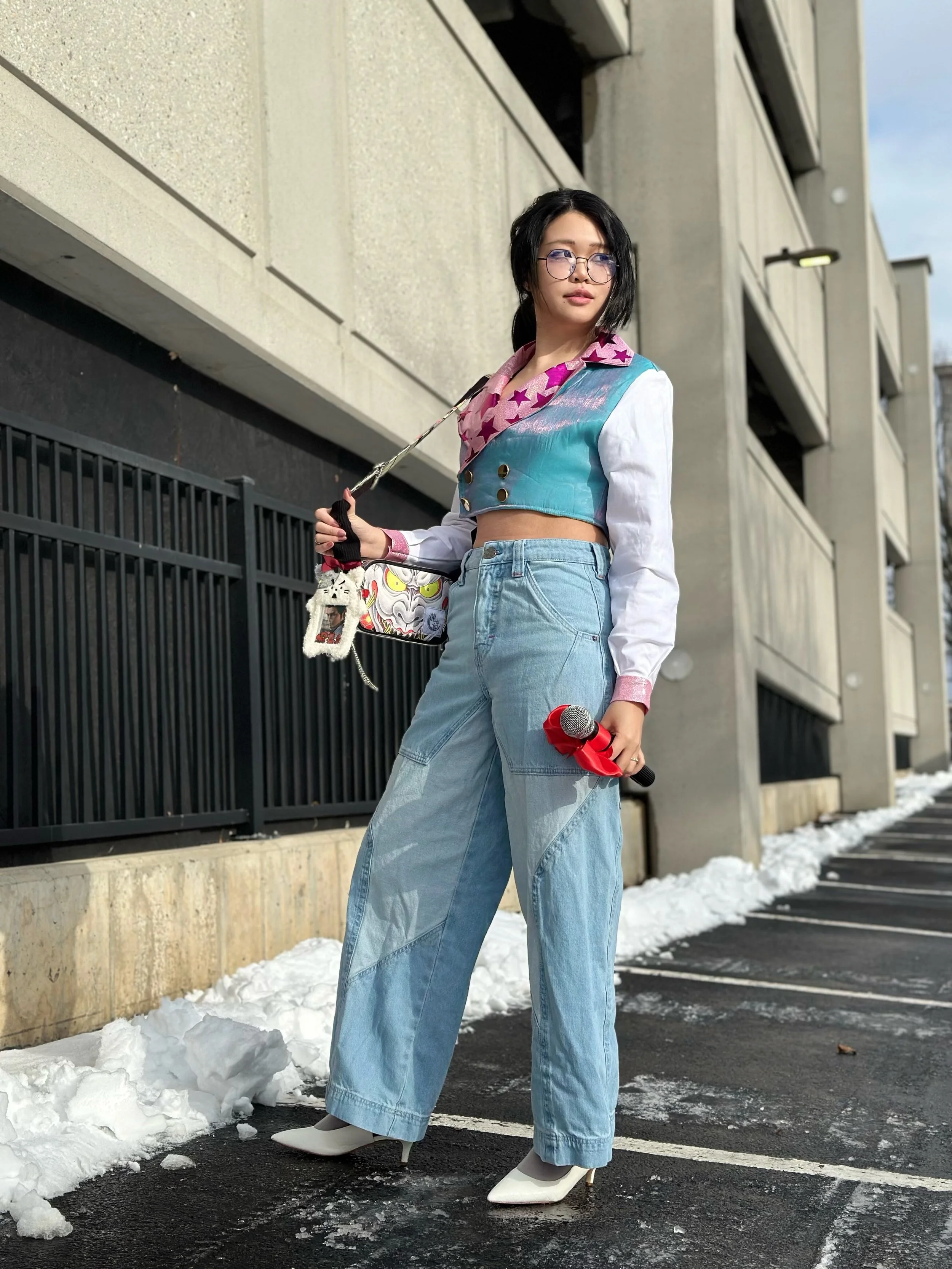 A young woman with black hair, glasses, and fashionable clothing standing outside in a parking lot, holding a microphone and a small bag, with snow on the ground.