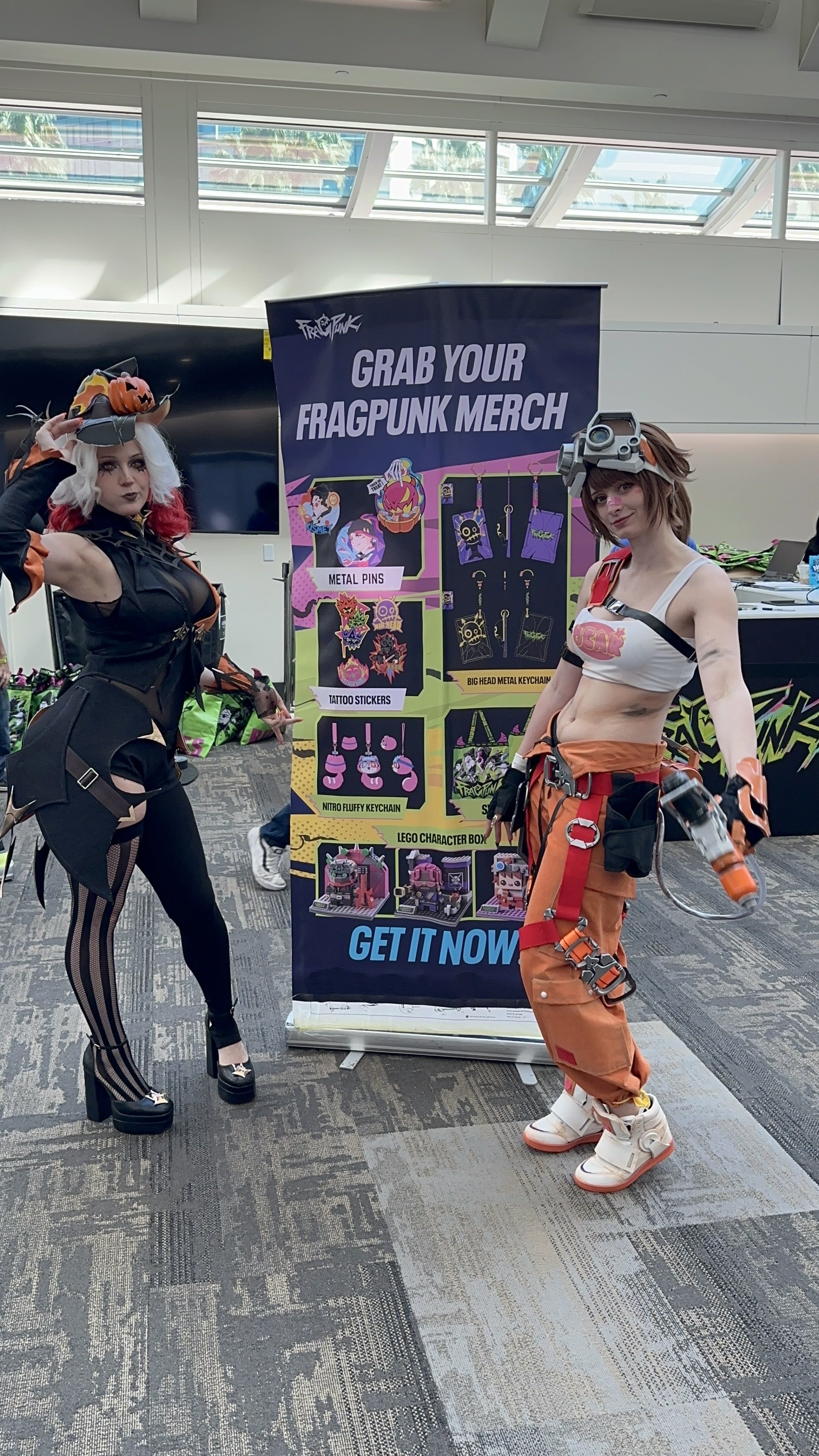 Two women in cosplay costumes at a convention, standing next to a colorful booth advertising FragPink merchandise. The woman on the left is dressed as a goth-themed character with a pumpkin hat, black and orange outfit, and striped stockings. The wom