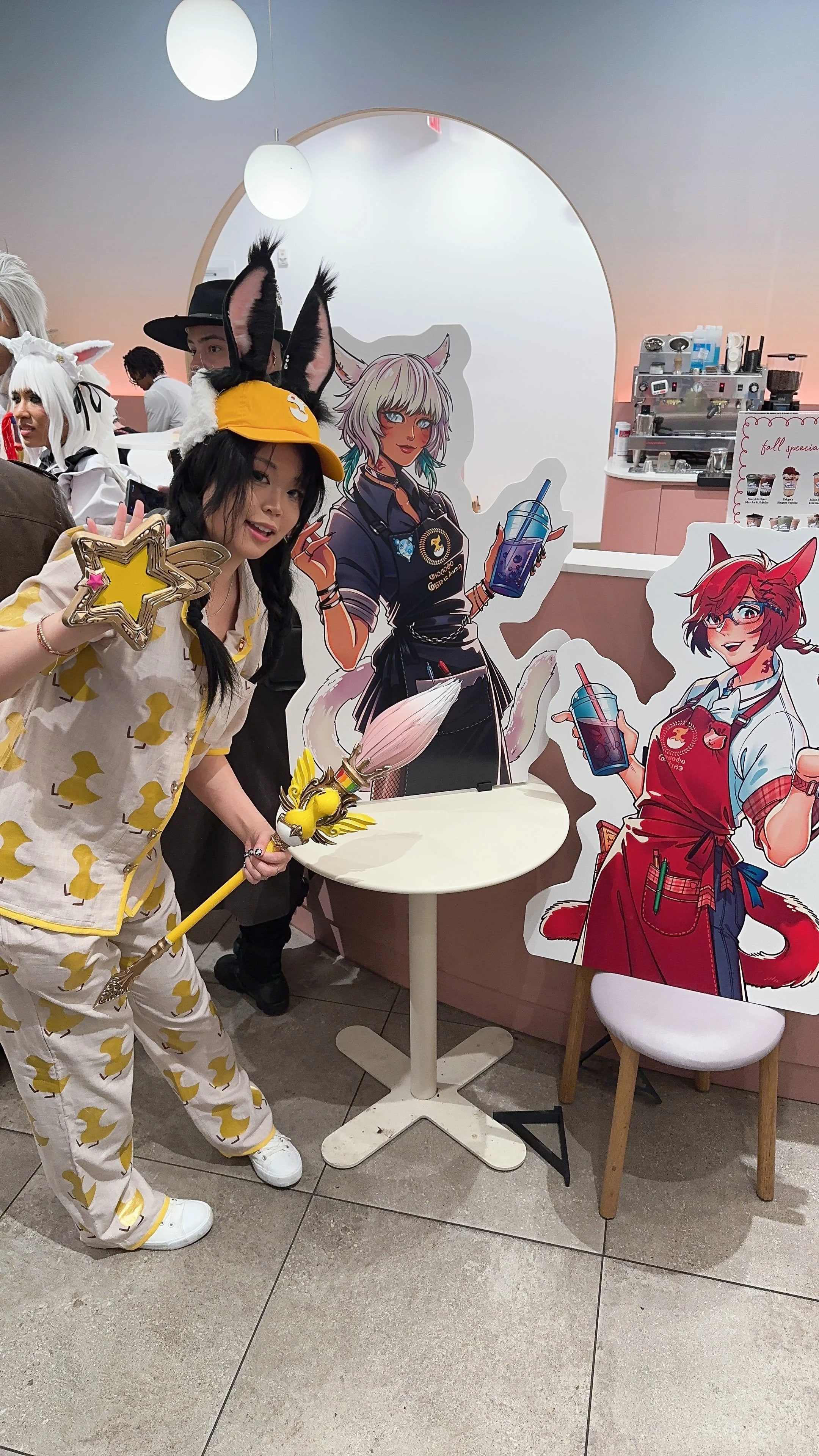 A woman dressed in a yellow and white duck-themed pajama set, wearing bunny ears and a yellow hat, holding a star-shaped wand, standing in front of colorful anime-style character cutouts in a cafe.