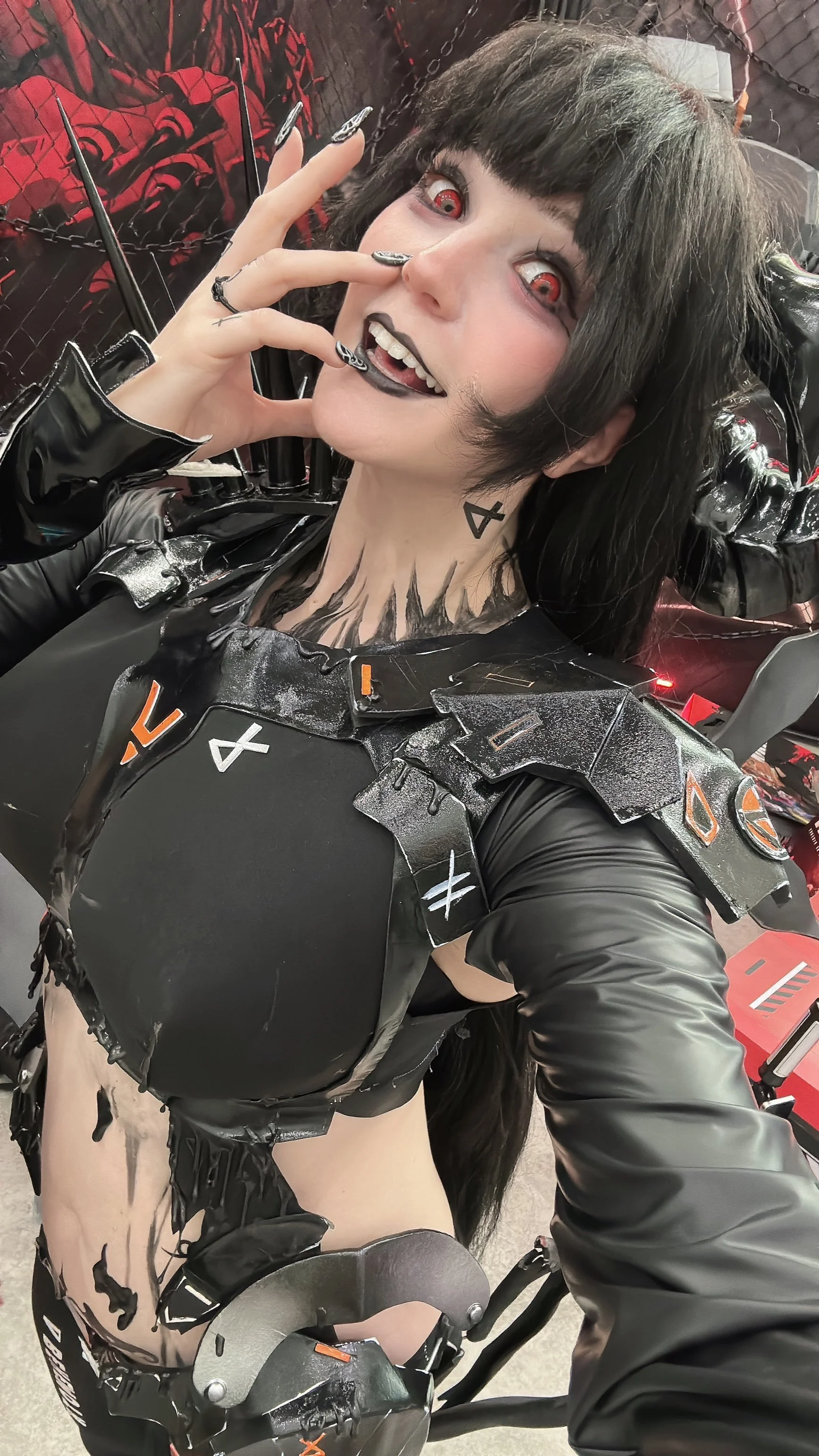 Person dressed in black with gothic style makeup, red contact lenses, and zombie makeup, taking a selfie in an indoor setting.