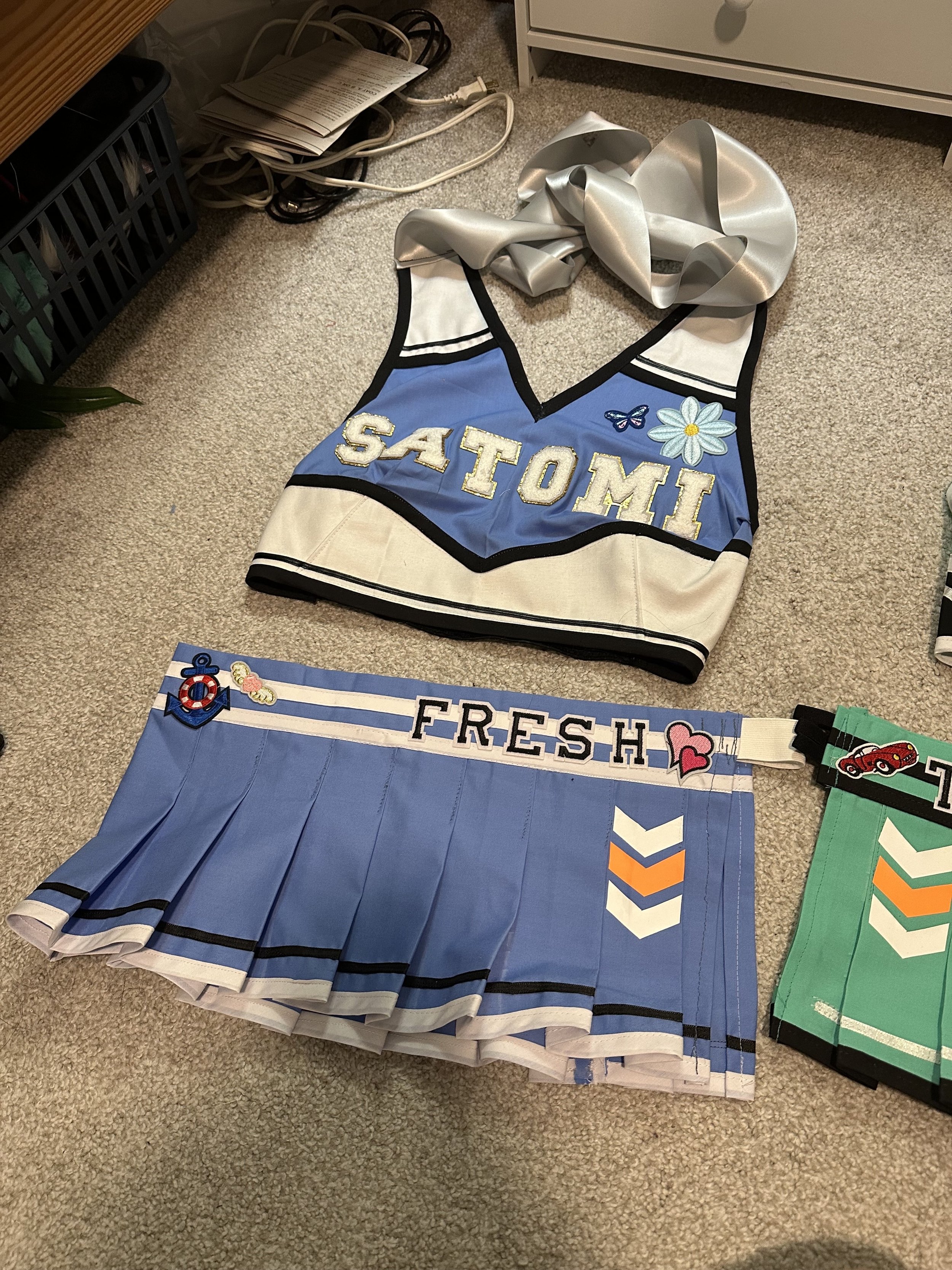 Cheerleading top with 'SATOMI' written on it and a floral and butterfly design, blue pleated skirt with 'FRESH' written and decorative patches, and another green pleated skirt in the background.