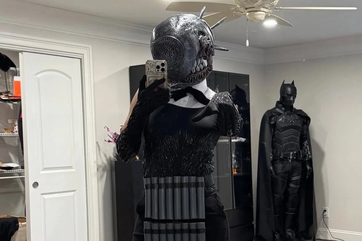 Person in a costume with a mask resembling a speaker, taking a mirror selfie in a room with superhero costumes, including a Batman figure, in the background.