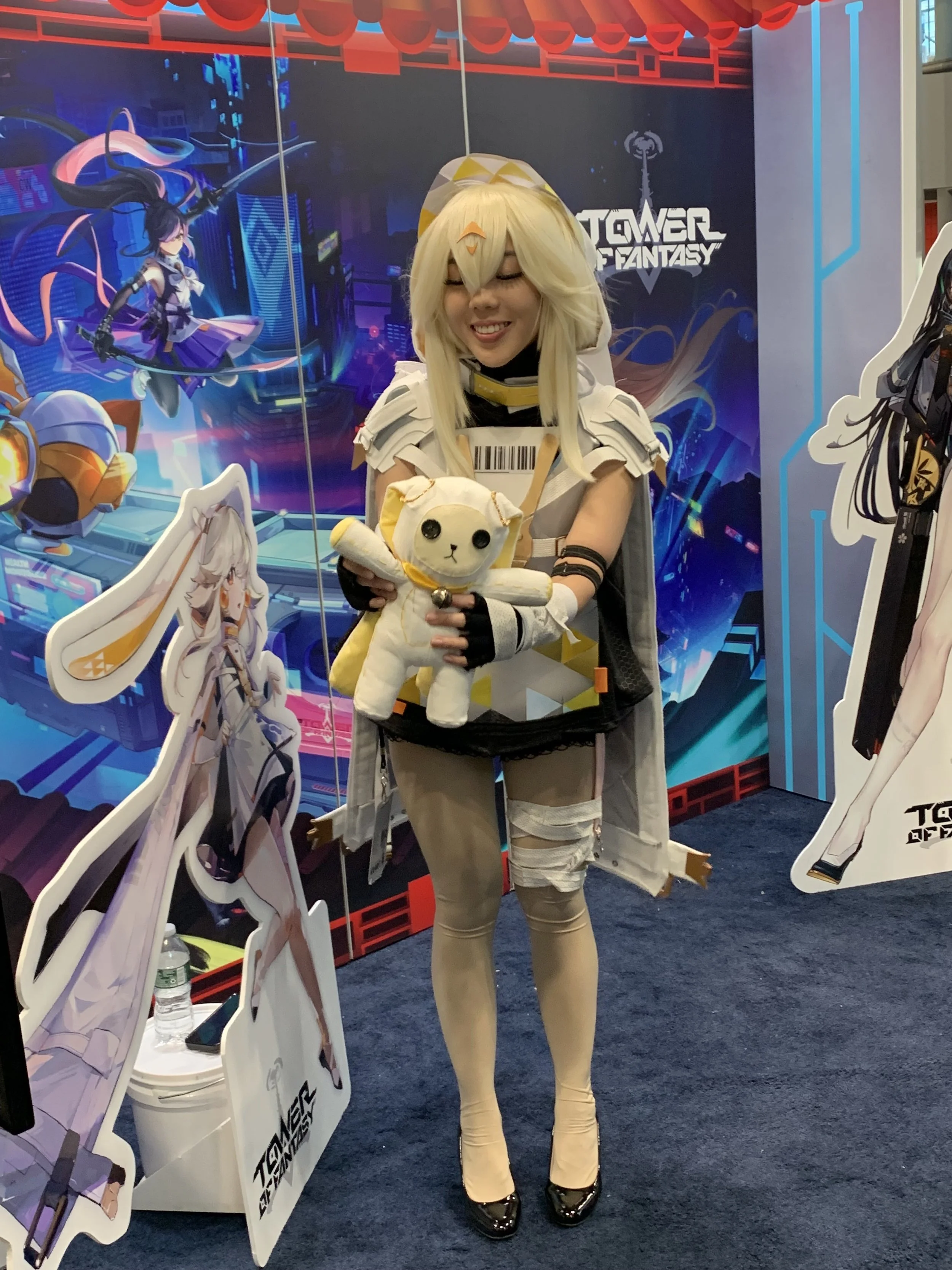 A woman dressed in a cosplay costume holding a plush toy in front of a colorful video game themed backdrop at a convention. The woman has blonde hair, a futuristic outfit, and black high heels.