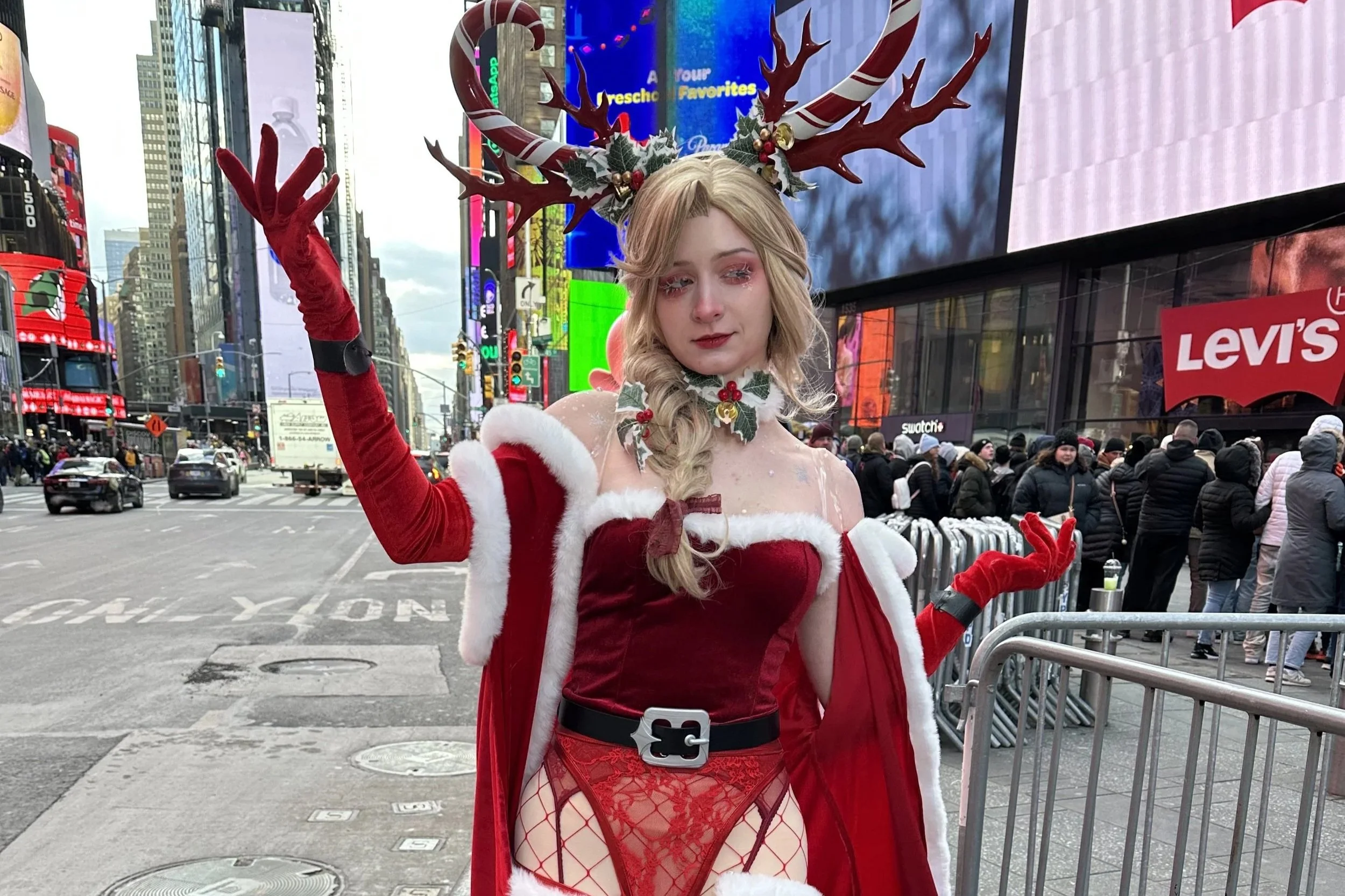 A woman dressed in a festive Santa-themed costume standing on a city street, wearing reindeer antlers, red gloves, and lace lingerie, with a crowd and tall buildings in the background.