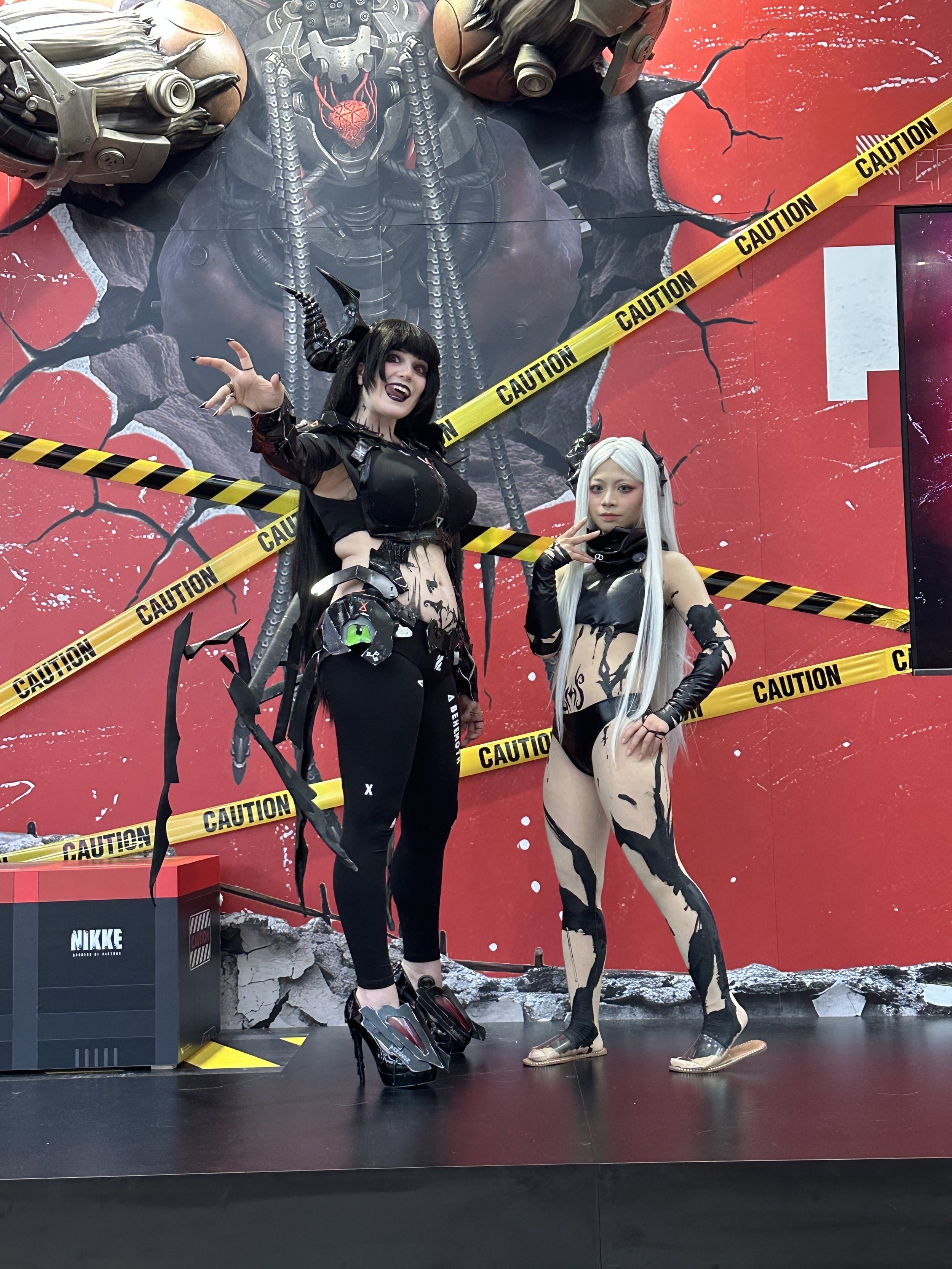 Two women dressed in black and white cosplay costumes, with dark and light hair, standing in front of a damaged red wall and a large poster of a character with a mechanical face and chains, surrounded by caution tape.