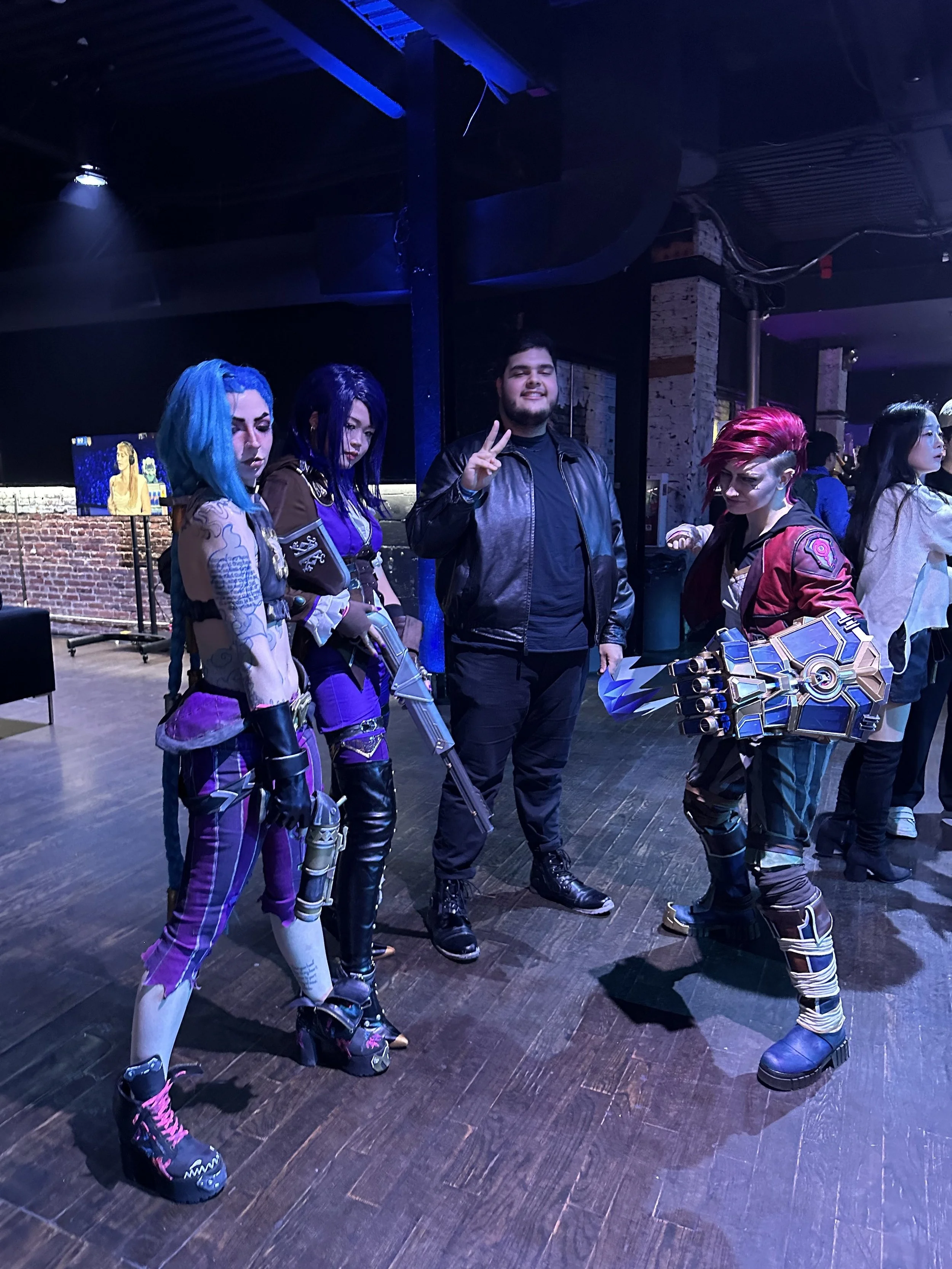 Group of five people, four women dressed in colorful, futuristic costumes with weapons, and one man in a black leather jacket making a peace sign, at an indoor event with dark lighting and exposed brick walls.