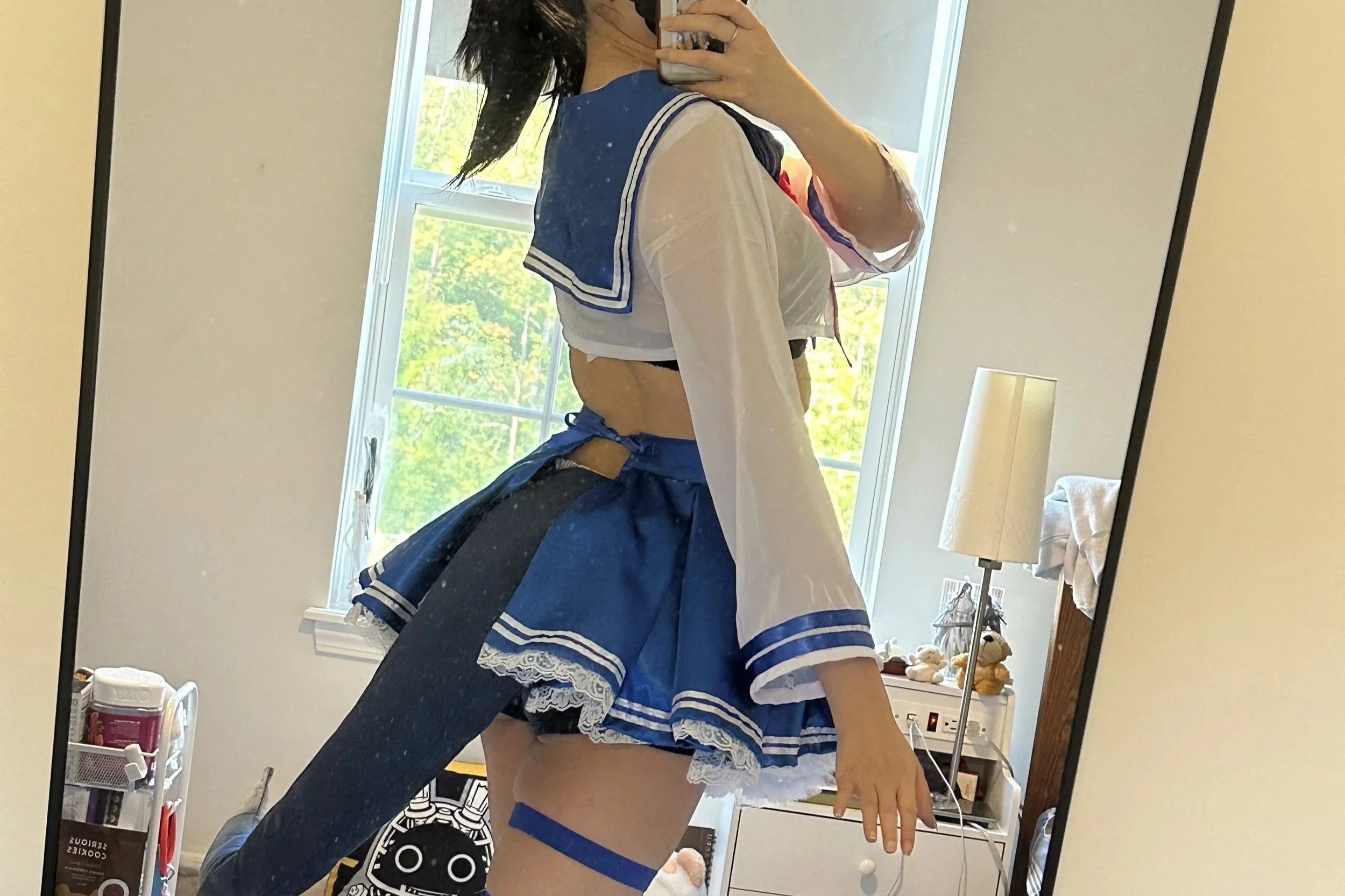 Person taking a mirror selfie dressed in a blue and white sailor-themed costume, with a bedroom background including a window, lamp, and various objects.