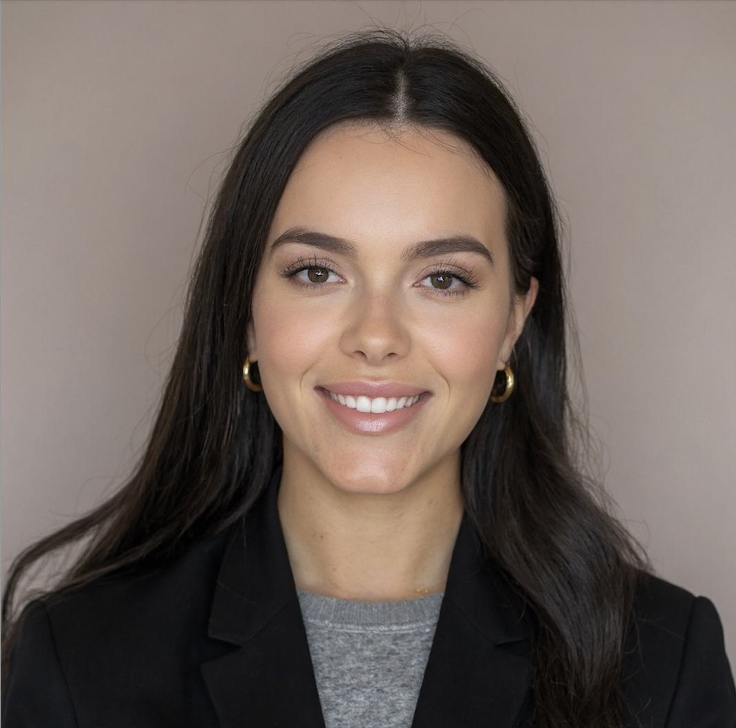 A woman with long dark hair, light skin, wearing a black blazer, gray top, and gold hoop earrings, smiling at the camera.