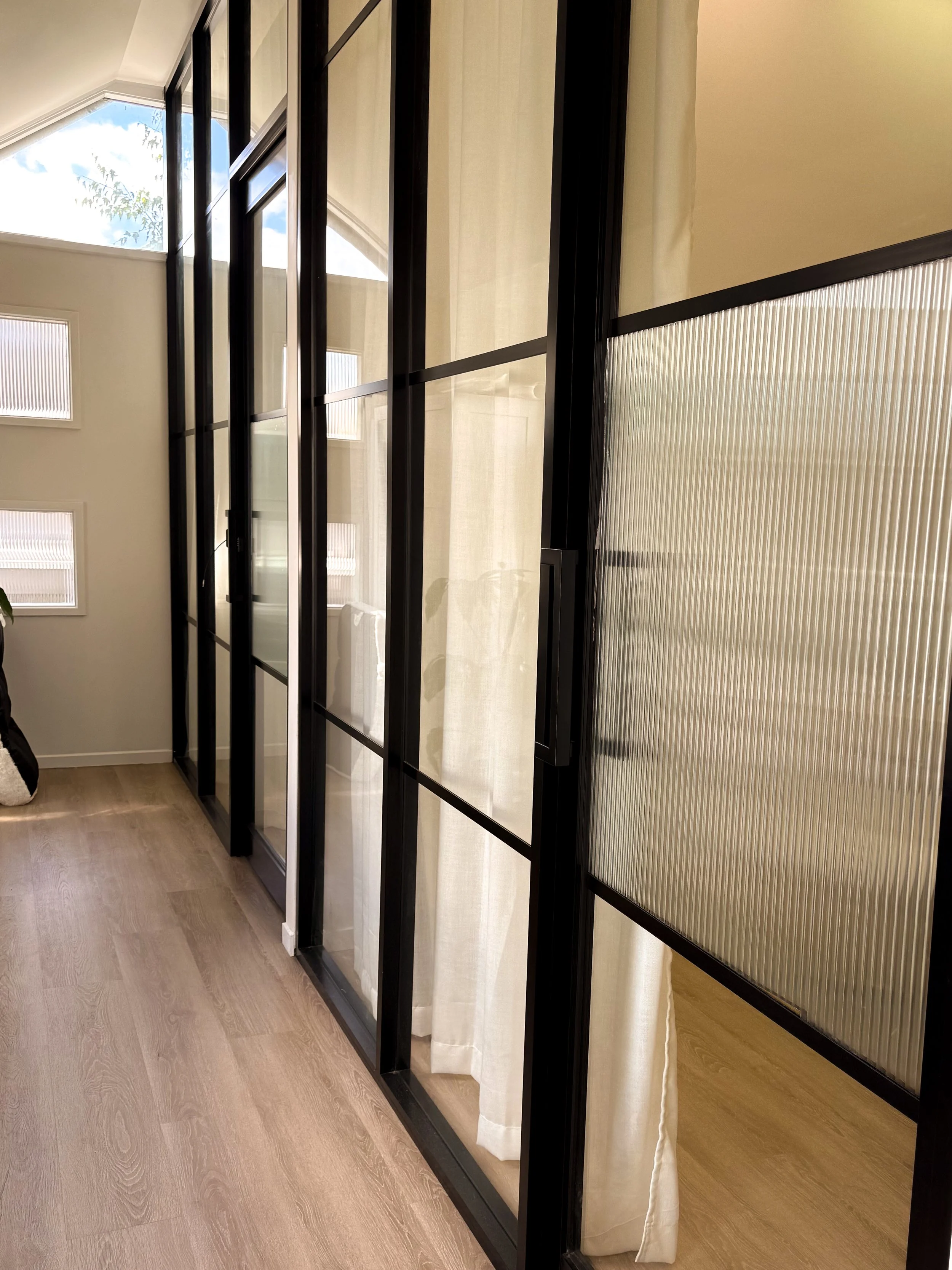 Glass partition with black metal framing and beige curtains inside a modern room with wood flooring, large windows, and a white ceiling.