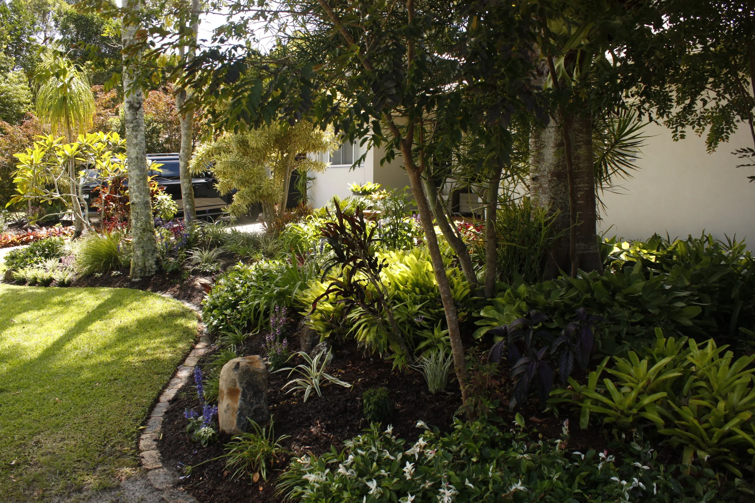 A lush, well-maintained garden with various green plants and trees, a curved brick border, and a paved driveway with a black vehicle parked in the background.
