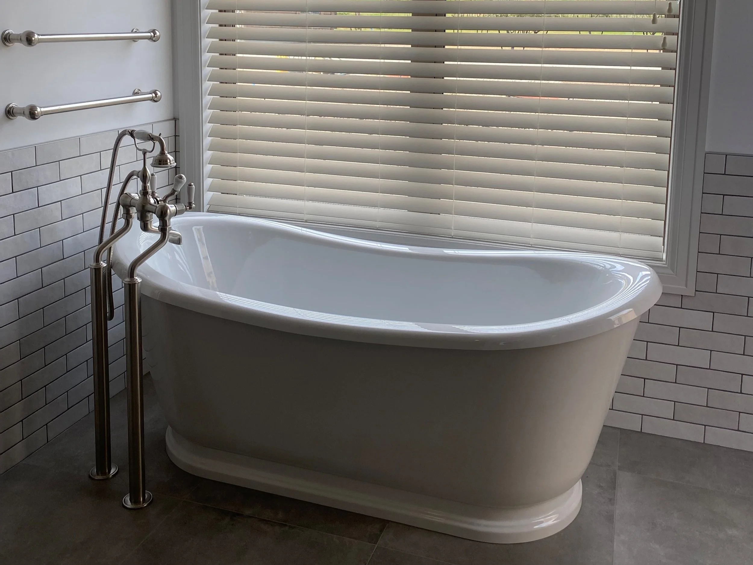 A white freestanding bathtub with a chrome faucet, situated in front of a window with beige blinds in a modern bathroom.