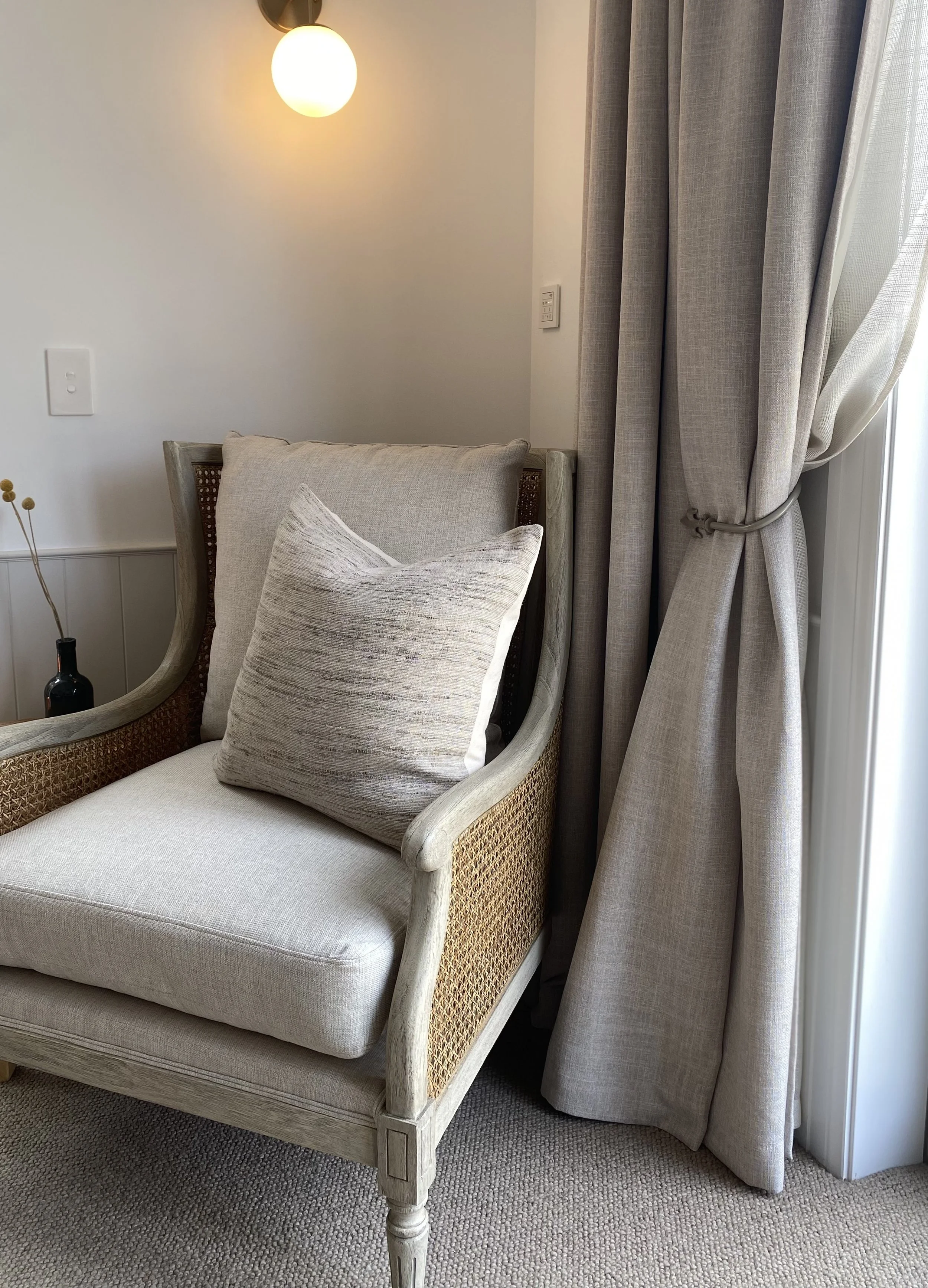 A beige armchair with a woven side panel and light beige cushions, placed next to a window with light gray curtains tied back, illuminated by a round wall-mounted light fixture.