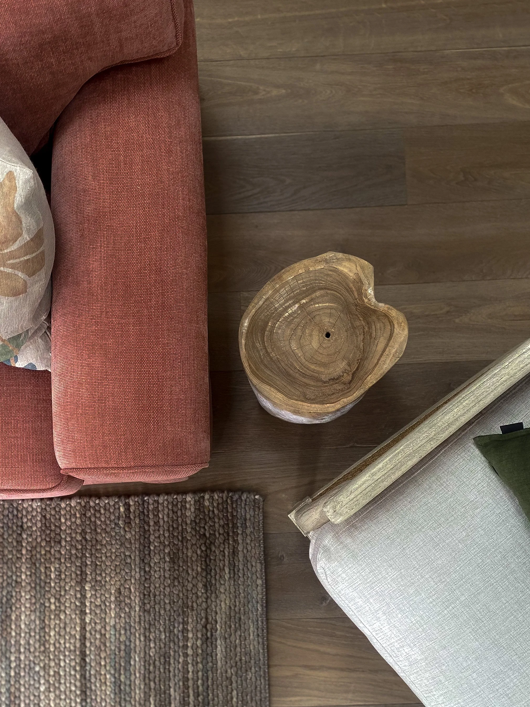 A circular wooden side table with a natural wood finish, placed on a wooden floor next to a red upholstered sofa with a floral pillow and a beige upholstered armchair with green fabric detail.