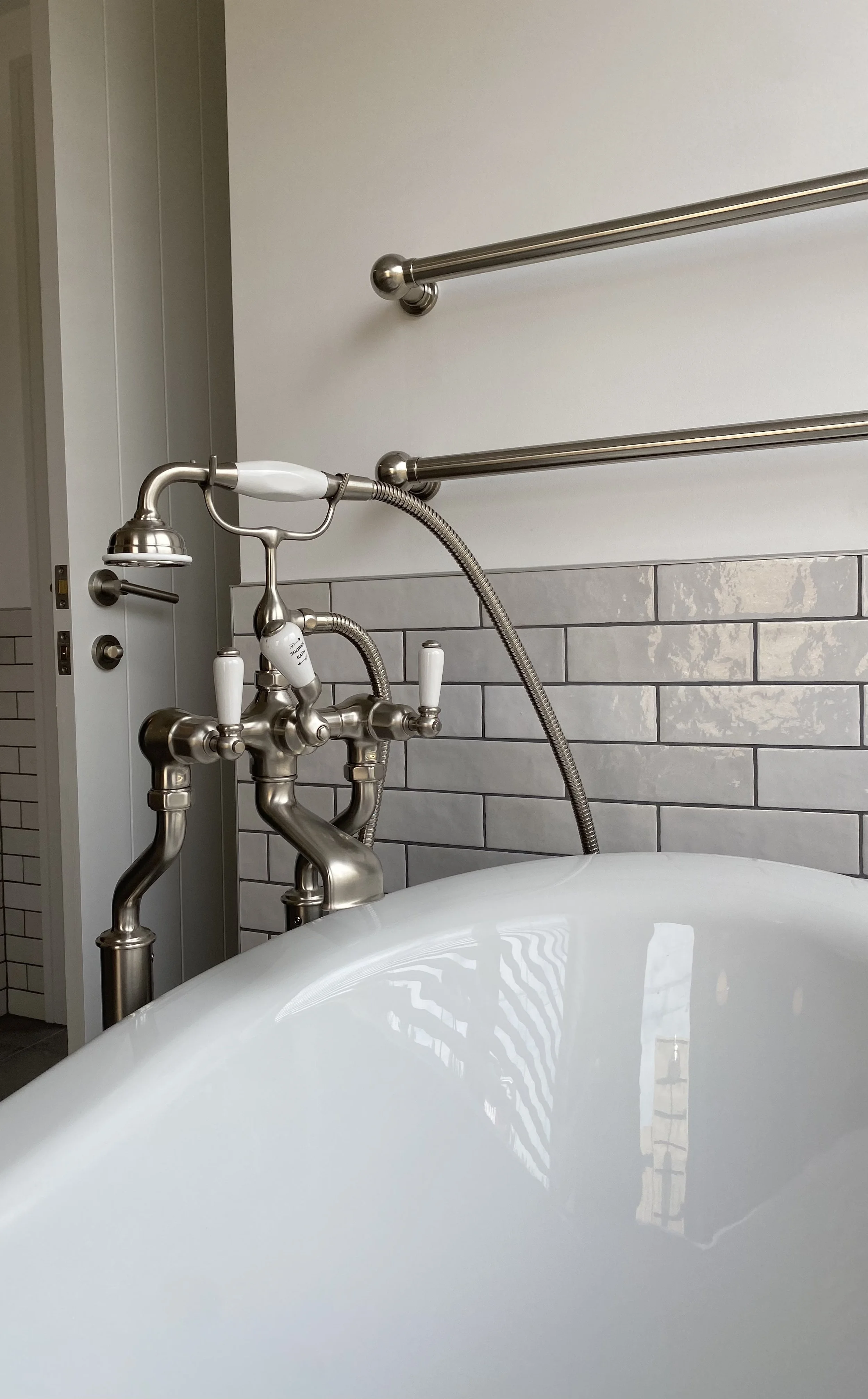 Close-up of a vintage-style bathtub faucet with handheld showerhead, chrome fixtures, and white handles, with a white tiled wall and a towel rack in the background.