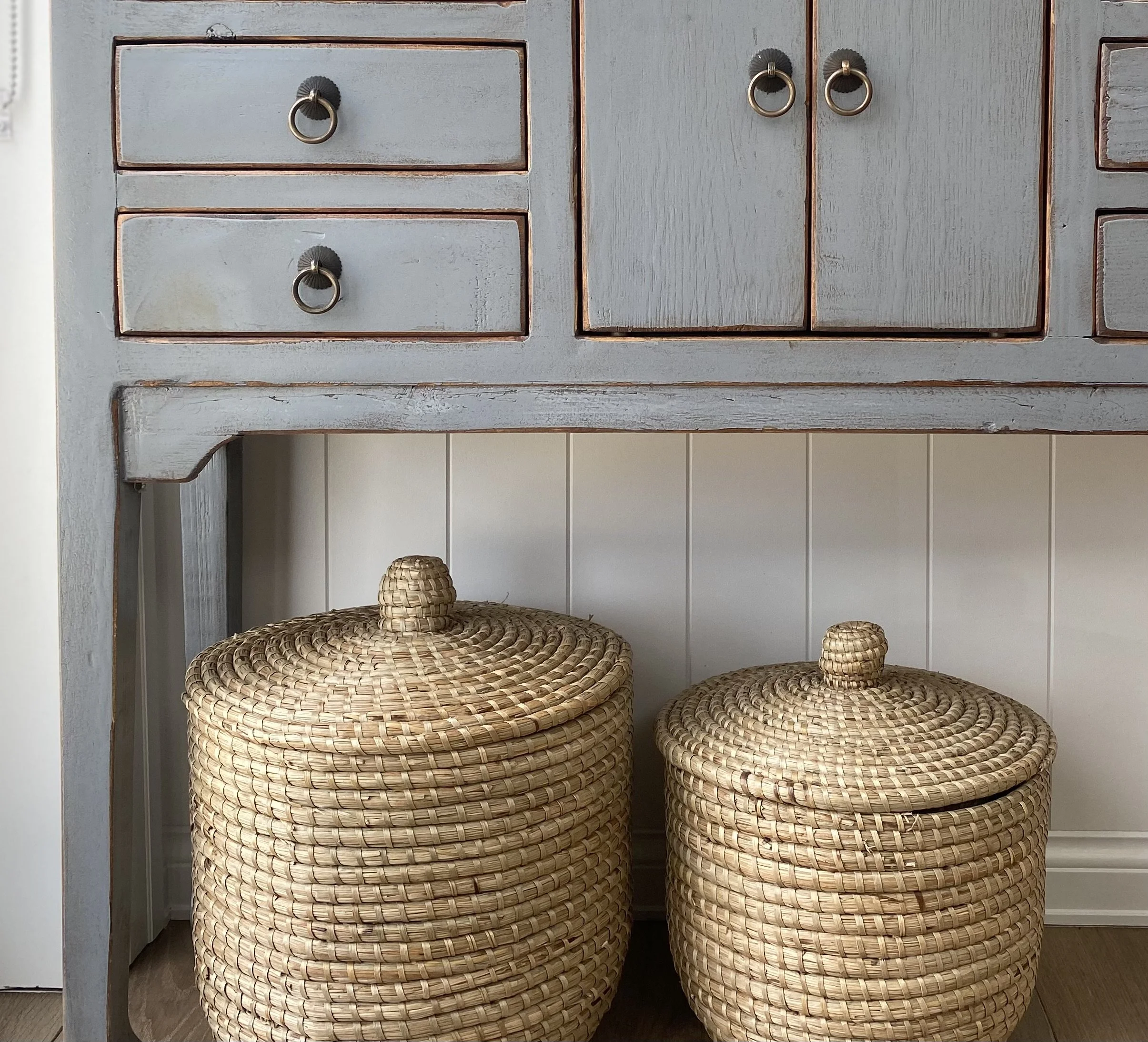Wooden cabinet painted light gray with metal ring pulls on the drawers and doors, standing above two round woven baskets with lids